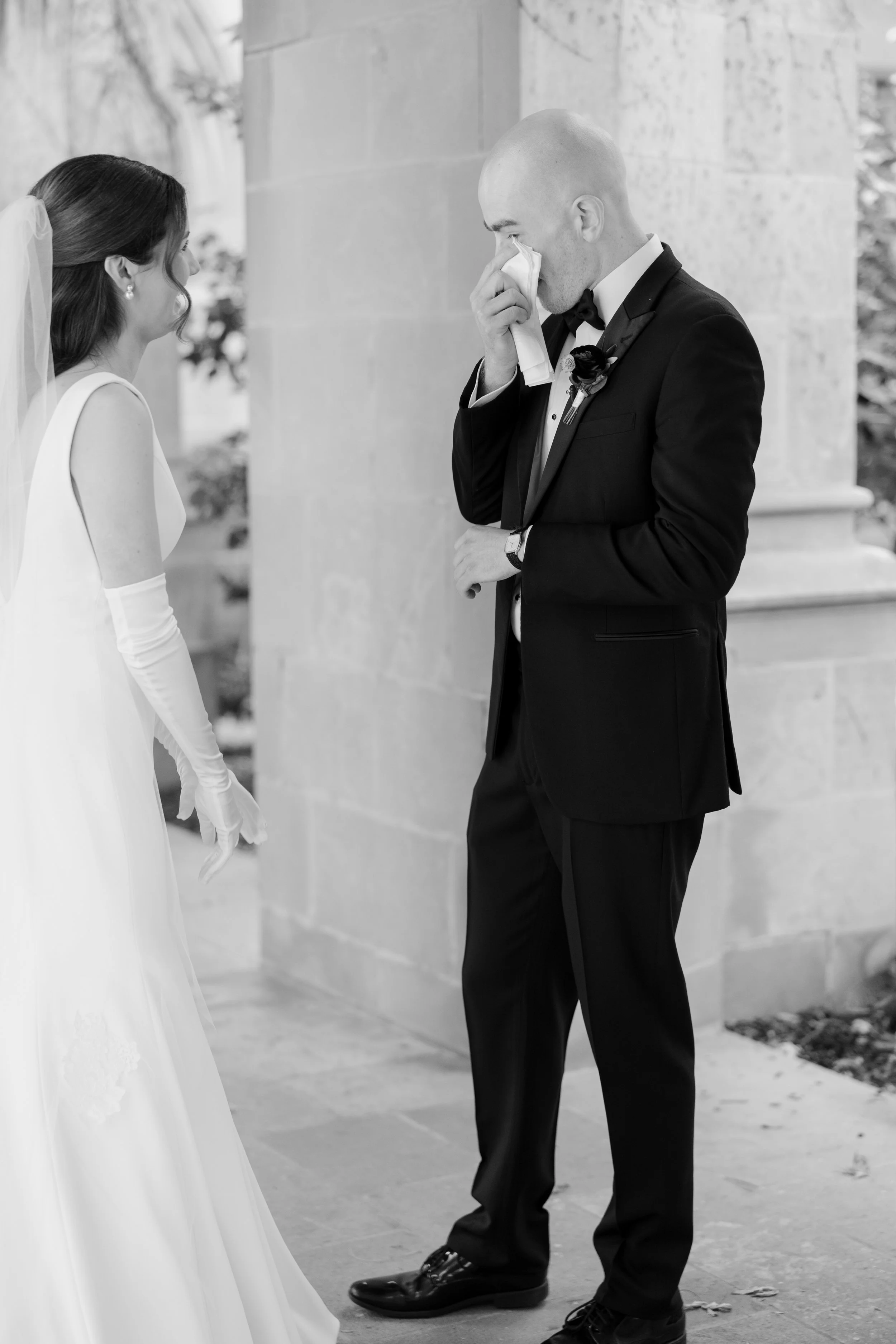 A man in a tuxedo wiping his eyes, possibly crying, while a woman in a wedding dress looks at him, standing outside on a stone walkway near a building.