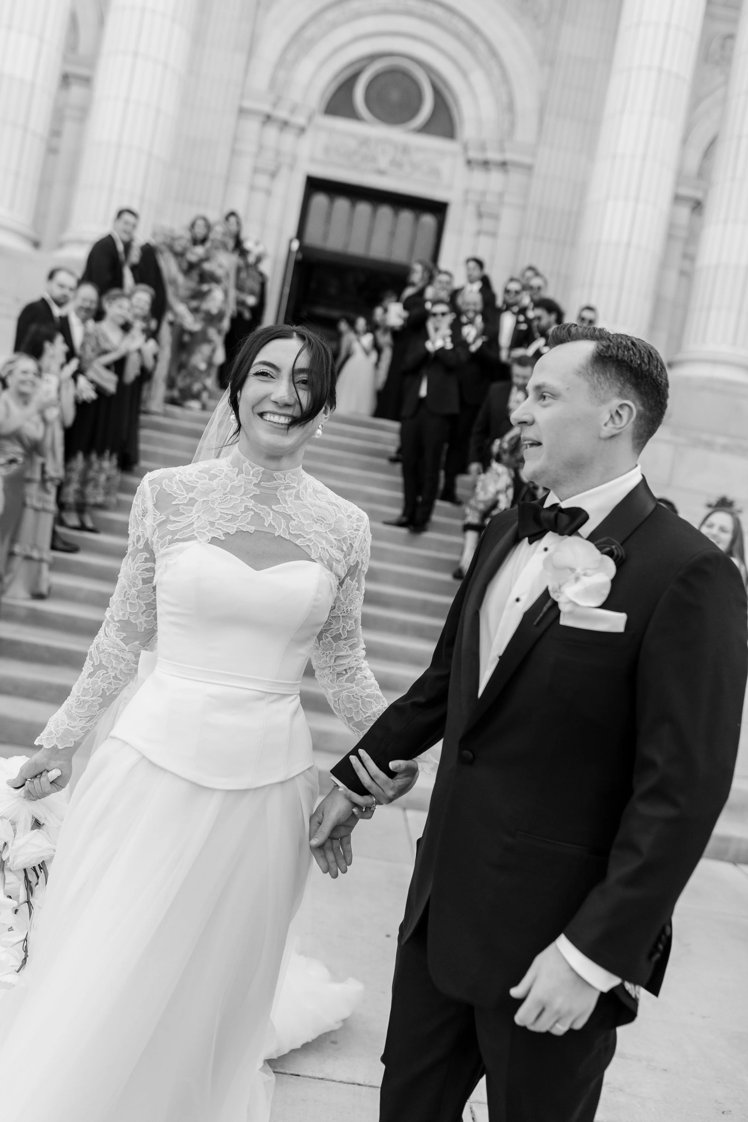 A bride and groom holding hands and smiling outside a church, surrounded by wedding guests.