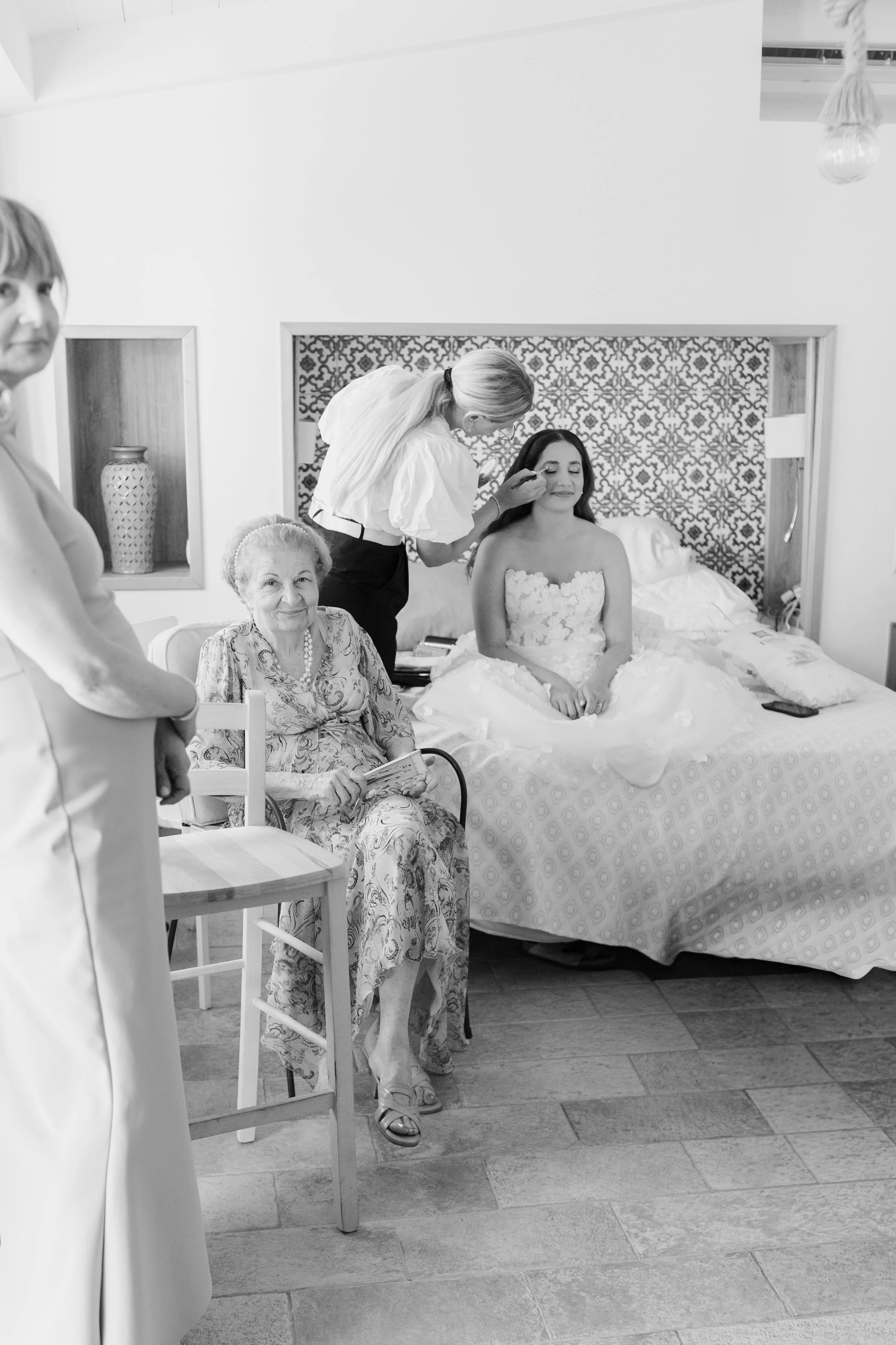 A bride in a strapless wedding dress sits on a bed while a makeup artist applies makeup to her face. An elderly woman with gray hair and a floral dress sits nearby, holding a book or magazine. The room has a patterned headboard and decorative vases o