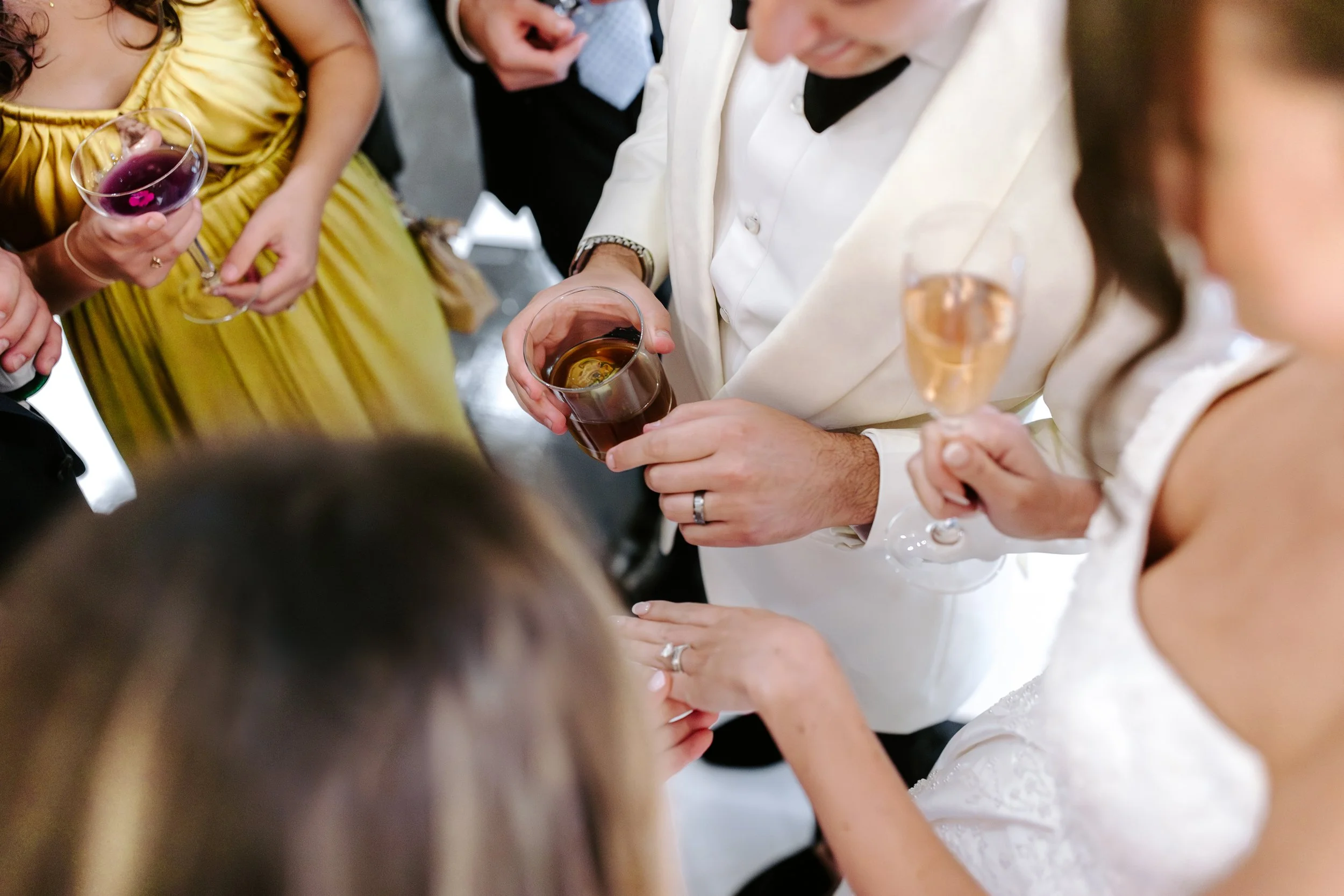 People at a social gathering holding glasses of wine and champagne, wearing formal attire, including a woman in a yellow dress and others in suits and dresses.