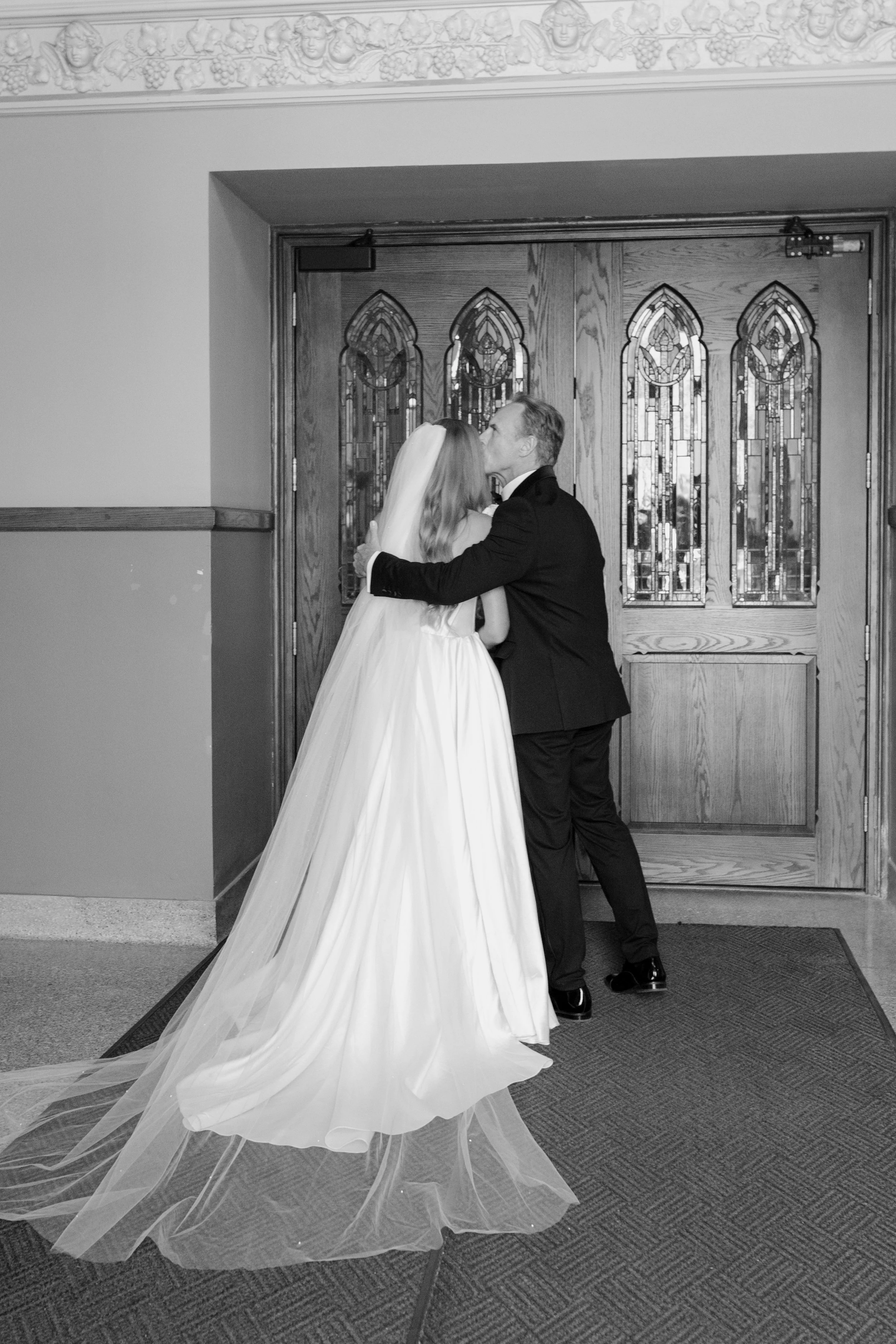 A bride and groom kiss in front of stained glass doors at their wedding. The bride wears a long white gown with a train, and the groom is dressed in a black suit.