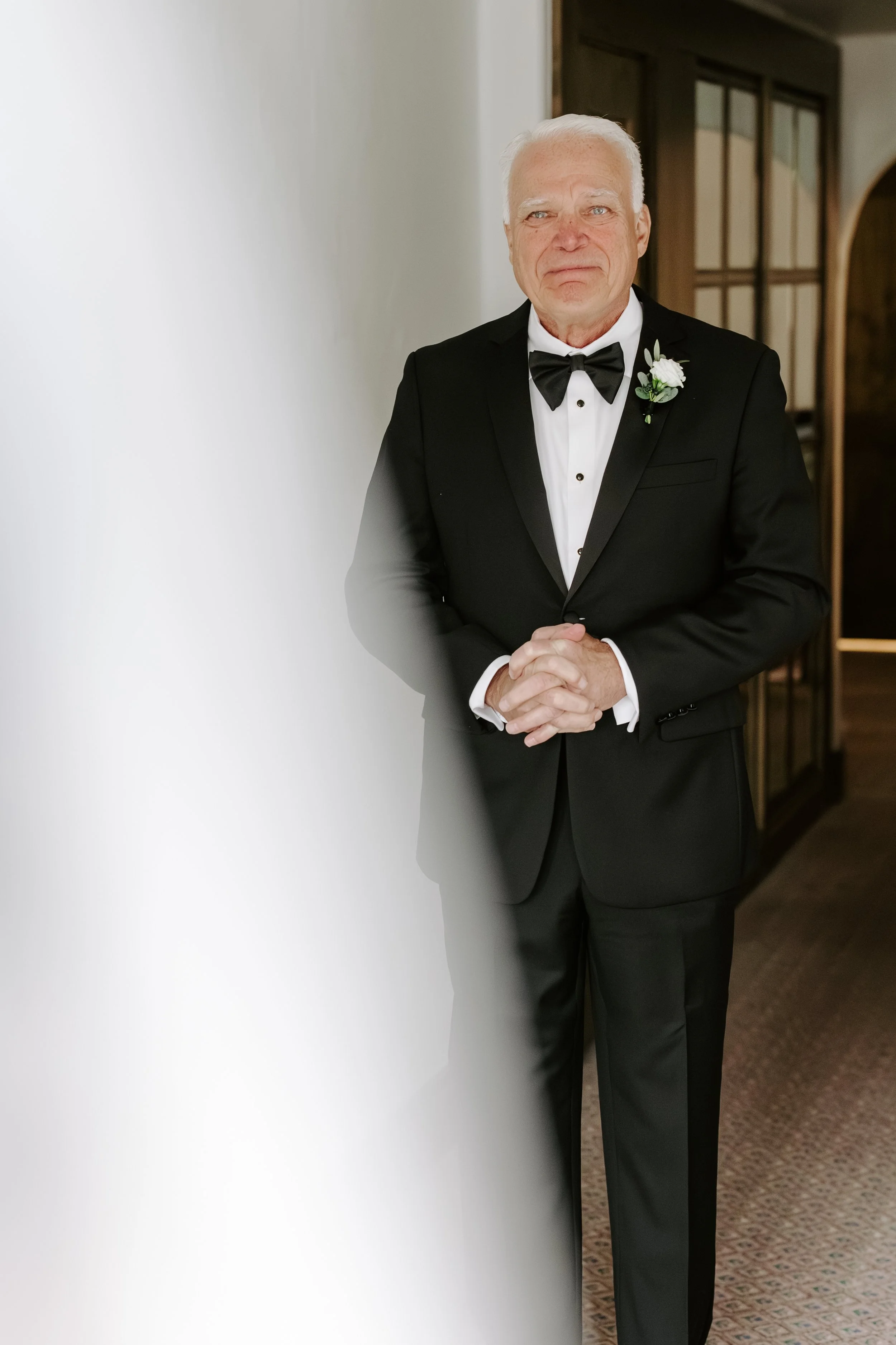 An elderly man in a black tuxedo with a bow tie and boutonniere, standing indoors and leaning against a white wall.