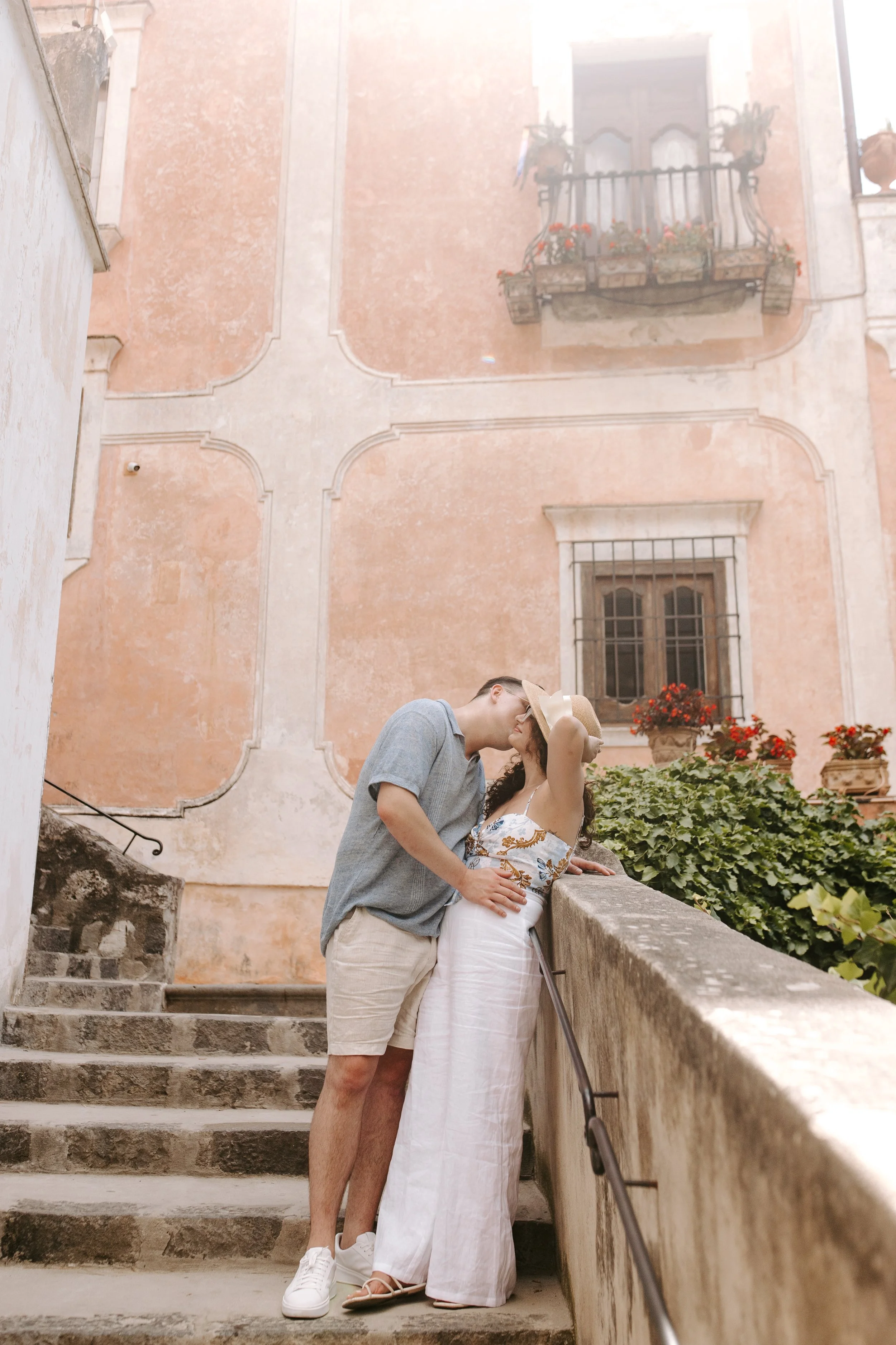 A couple kissing on a stone staircase in front of a pink historical building with flower boxes and ornate window.