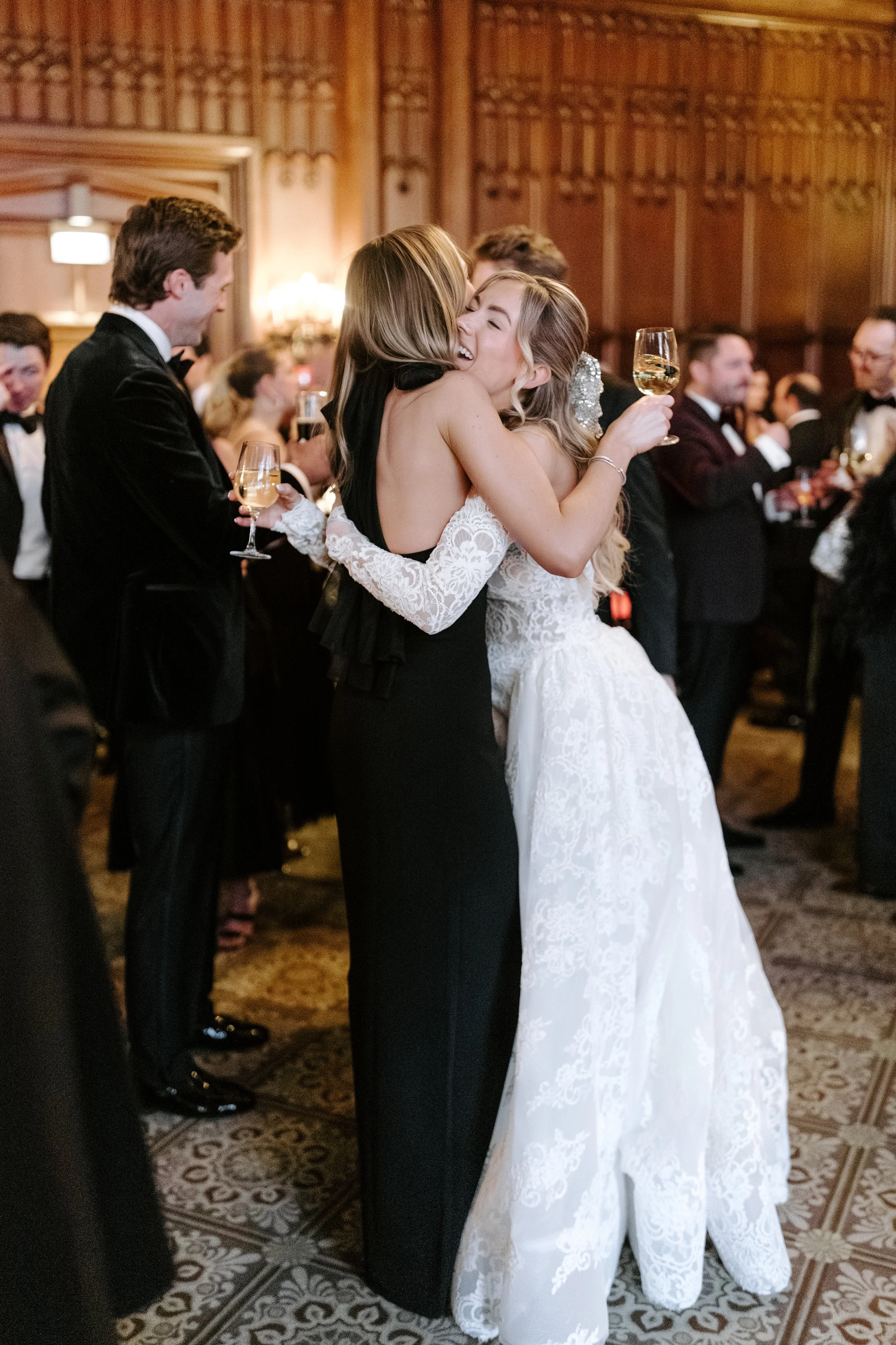 People at a wedding reception, embracing and holding glasses of white wine, in a warmly lit ornate wood-paneled room.