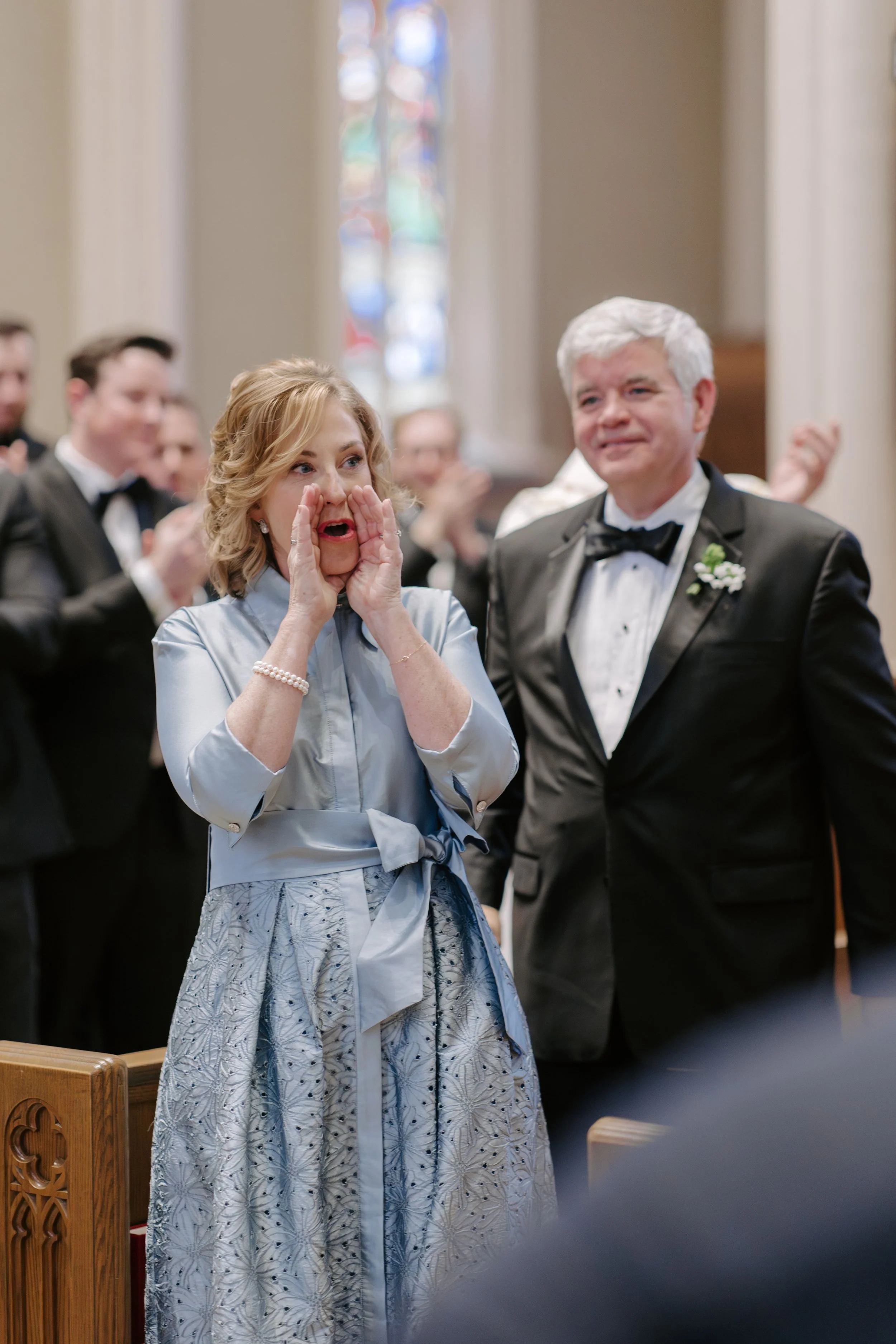 A woman in a light blue dress with a patterned skirt and pearl bracelet is loudly whispering into the ear of a man in a tuxedo with a boutonniere. A congregation of people in formal attire is clapping in the background inside a church with stained gl