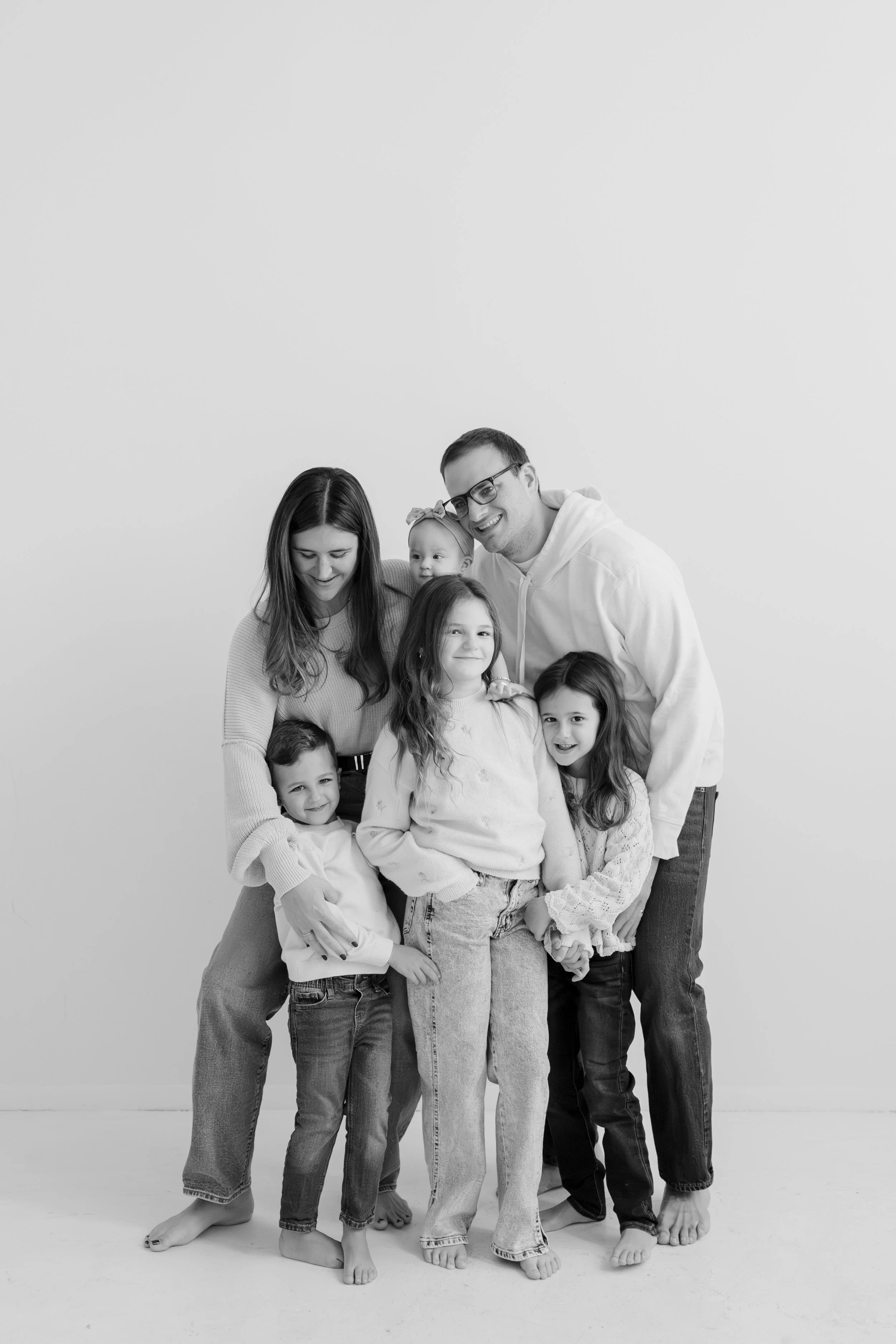 Black and white photo of a large family group with six children and two adults, all smiling and holding hands together, standing on a plain floor against a plain background.