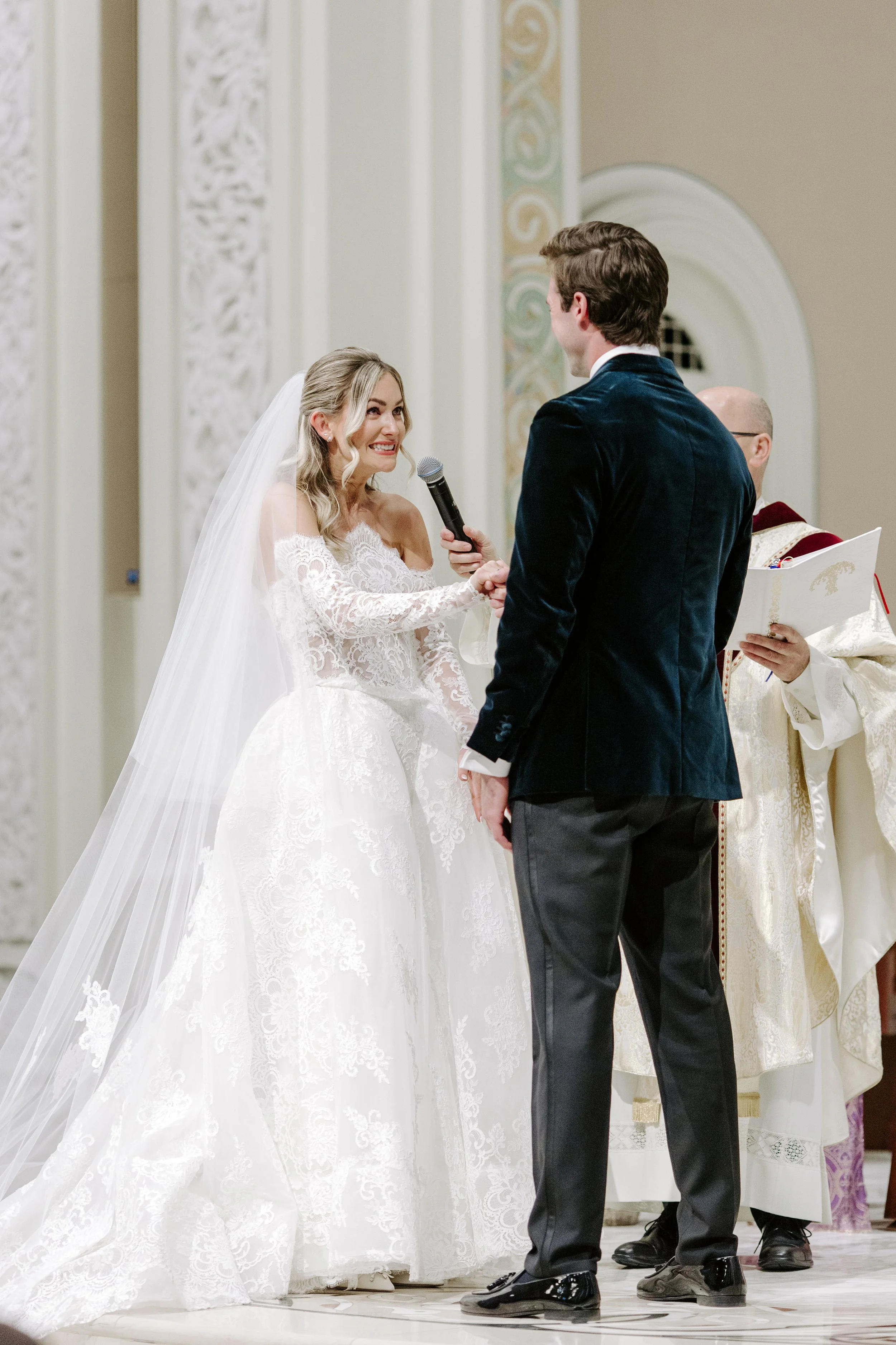 A bride and groom exchanging vows during a wedding ceremony in a church, with a priest officiating in the background.
