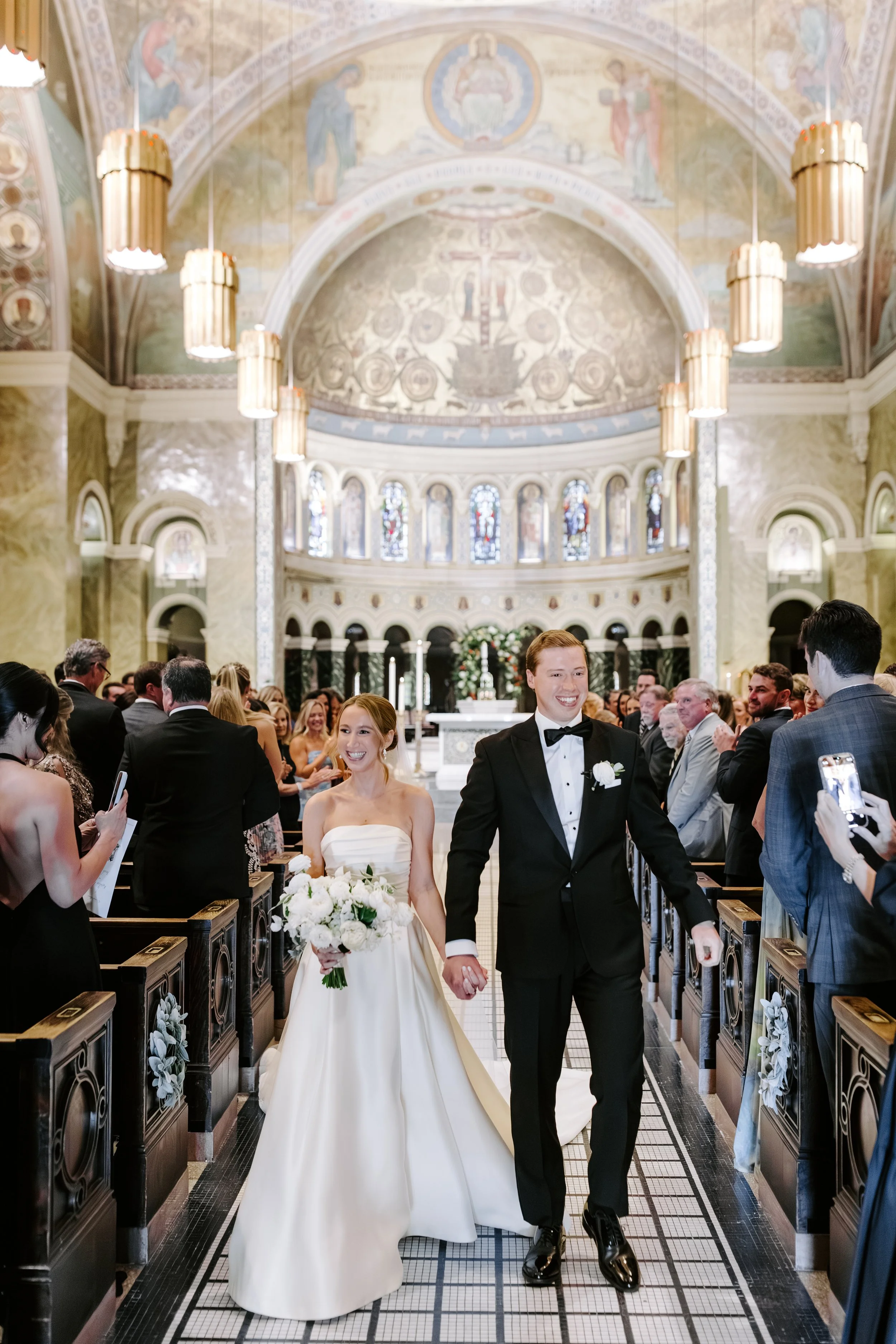 A bride and groom walking down the aisle of a church, smiling, holding hands, surrounded by guests. The church has a high, ornate ceiling with stained glass windows, chandeliers, and religious artwork.