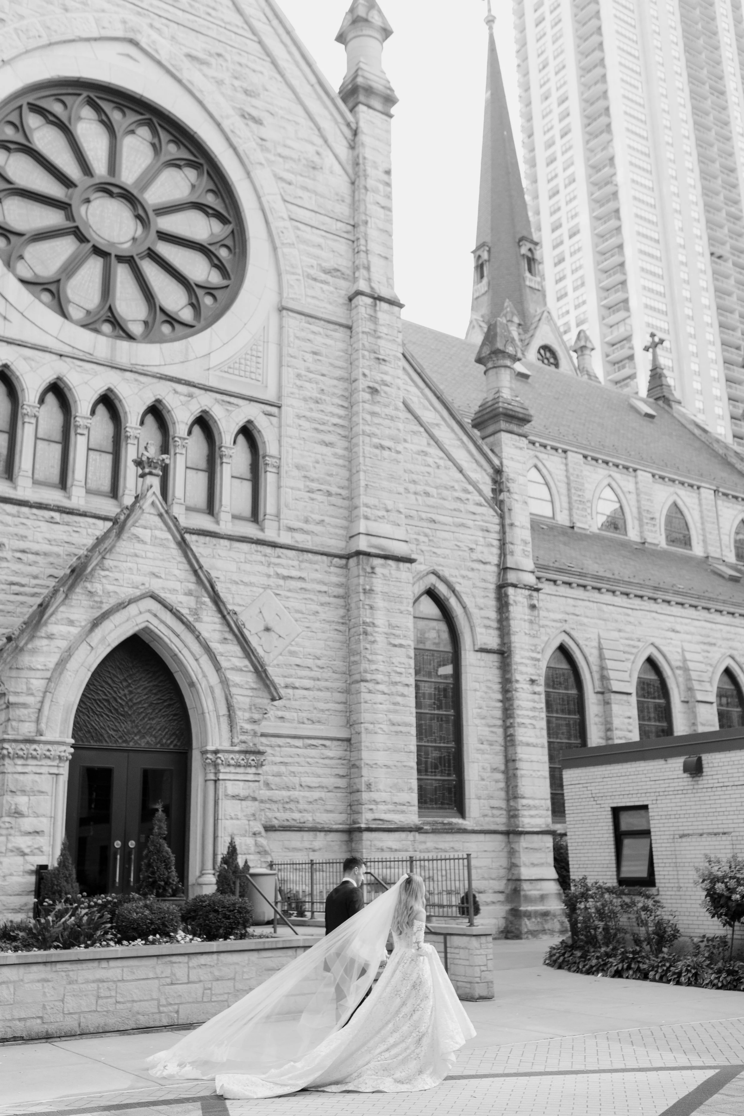 A bride in a wedding gown with a long train and veil and a groom in a tuxedo standing outside a stone church, with high-rise buildings in the background.