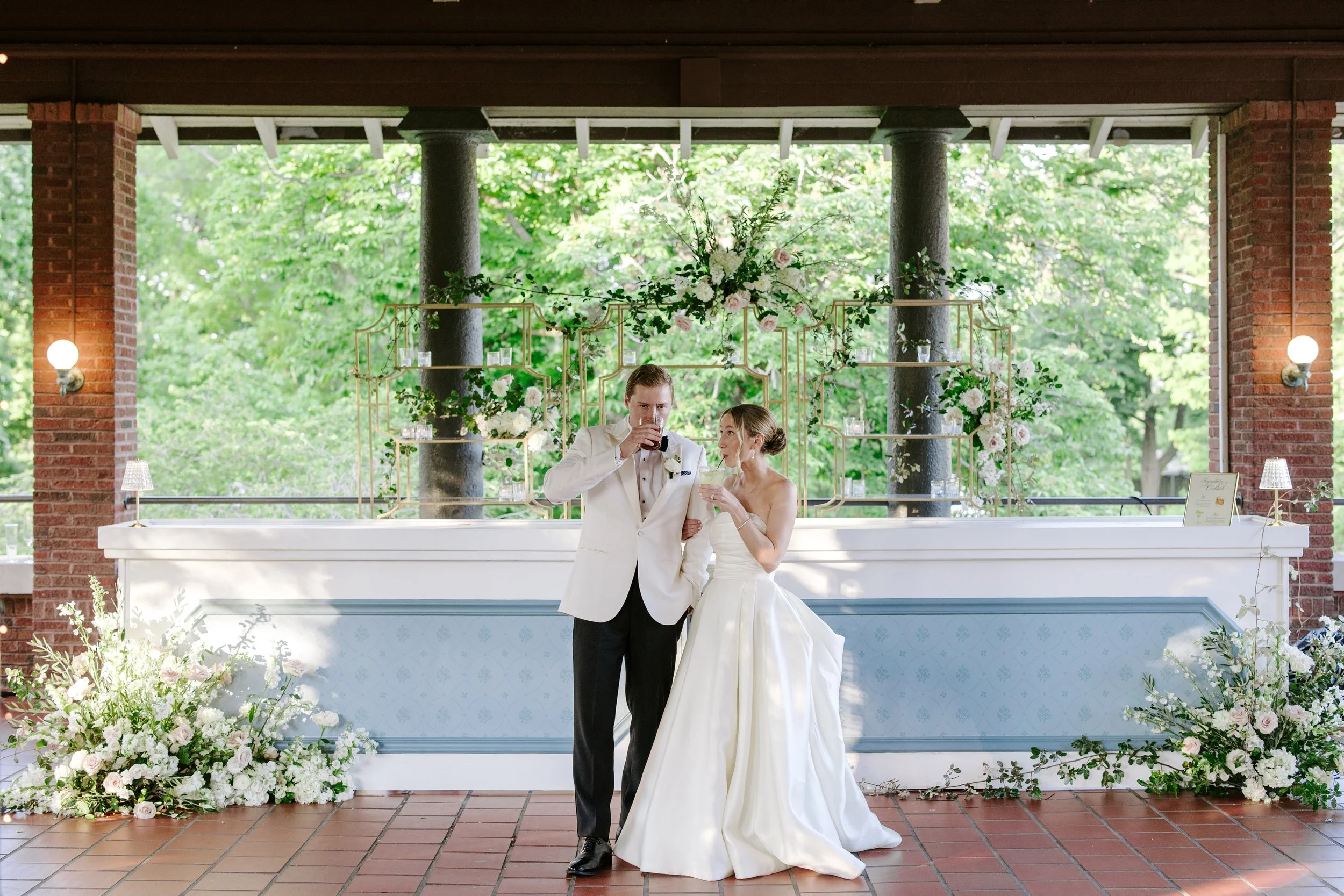 A bride and groom share a toast at their wedding reception outdoors, with green trees in the background and decorated floral arrangements on a table.