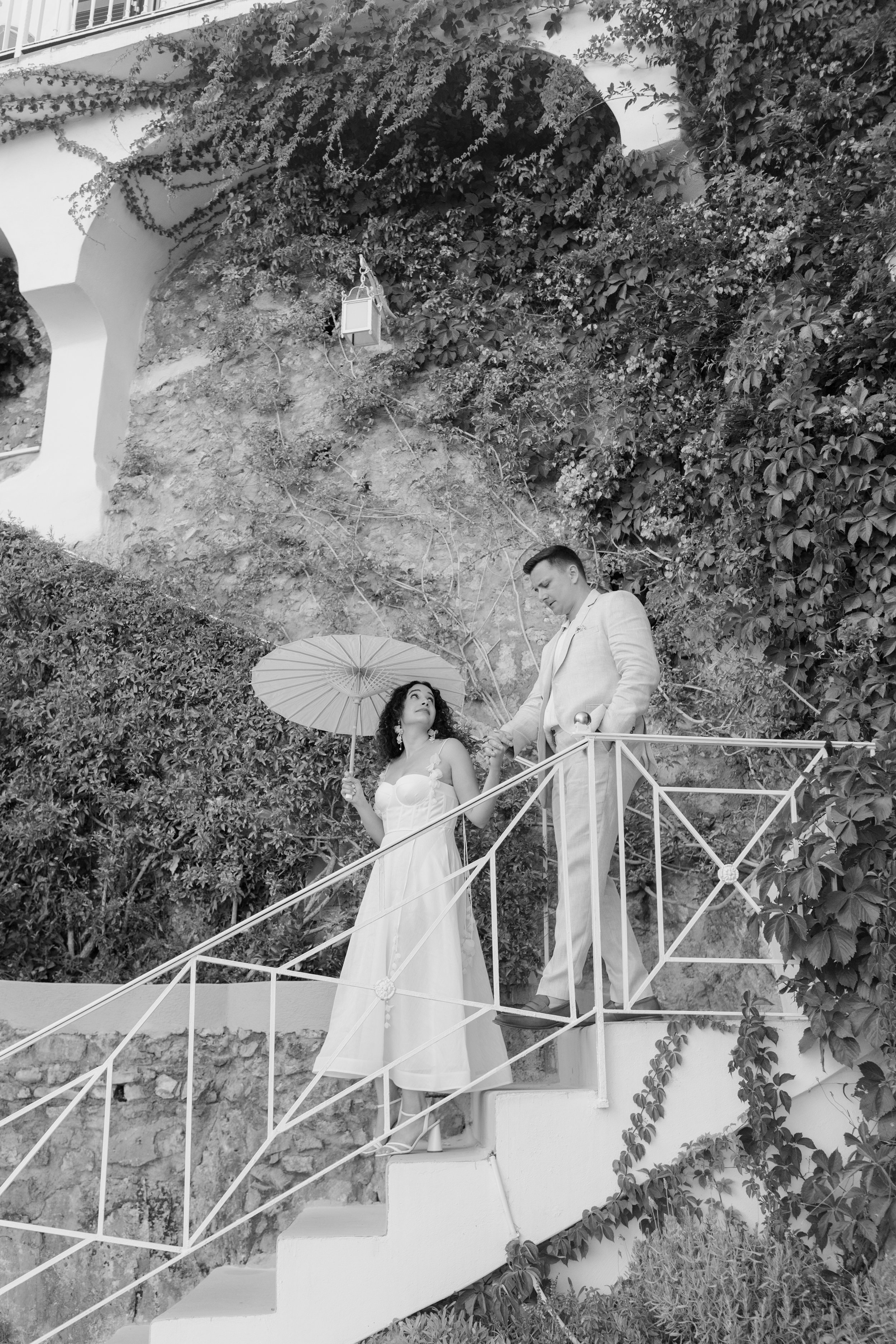 A black and white photo of a man and woman on a staircase outdoors. The woman is holding a parasol and looking up at the man, who is standing on the stairs and looking at her. The background includes a textured stone wall and lush foliage.
