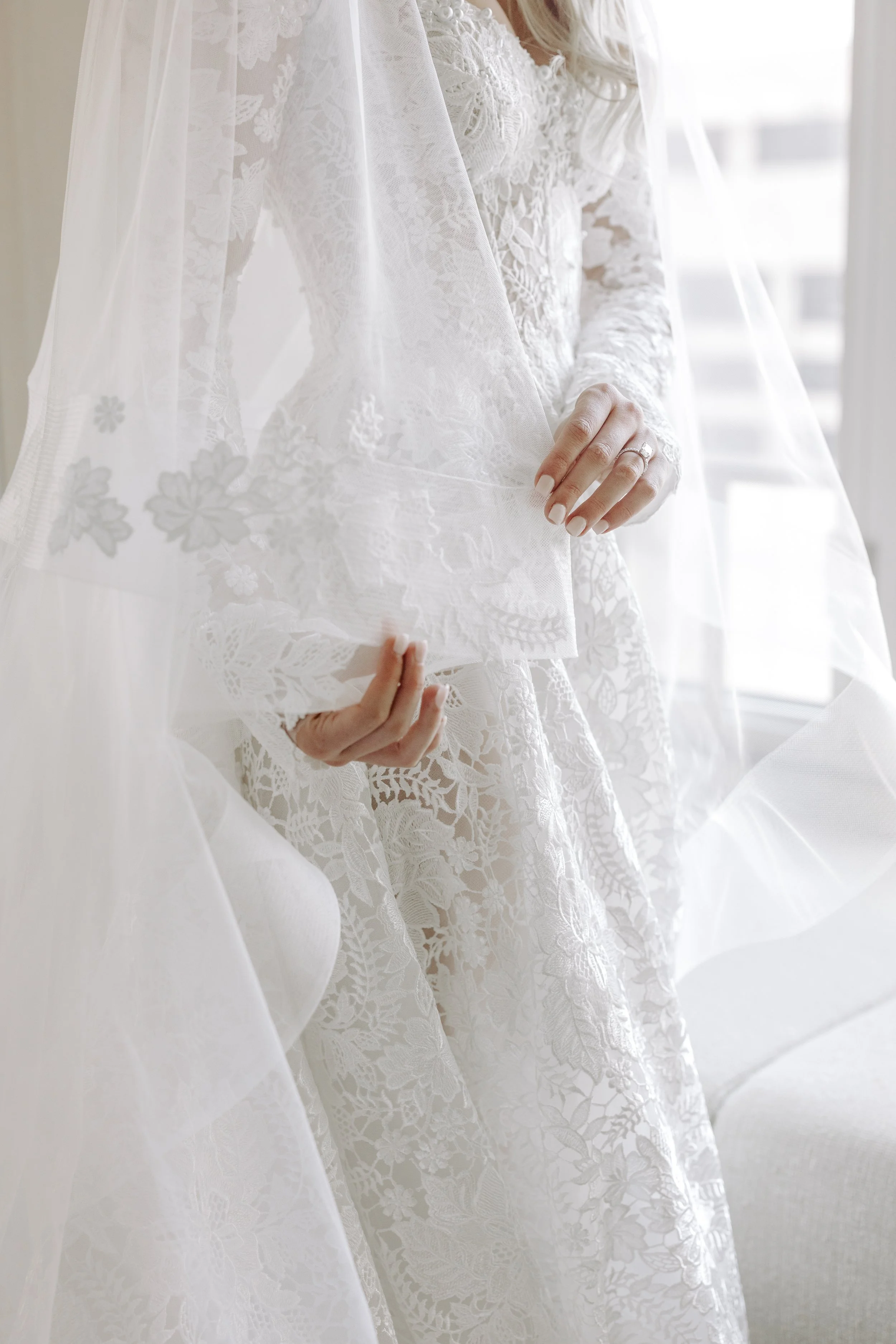 A woman wearing an elegant white lace wedding dress, partially covered by a sheer lace veil, standing near a window with sunlight.