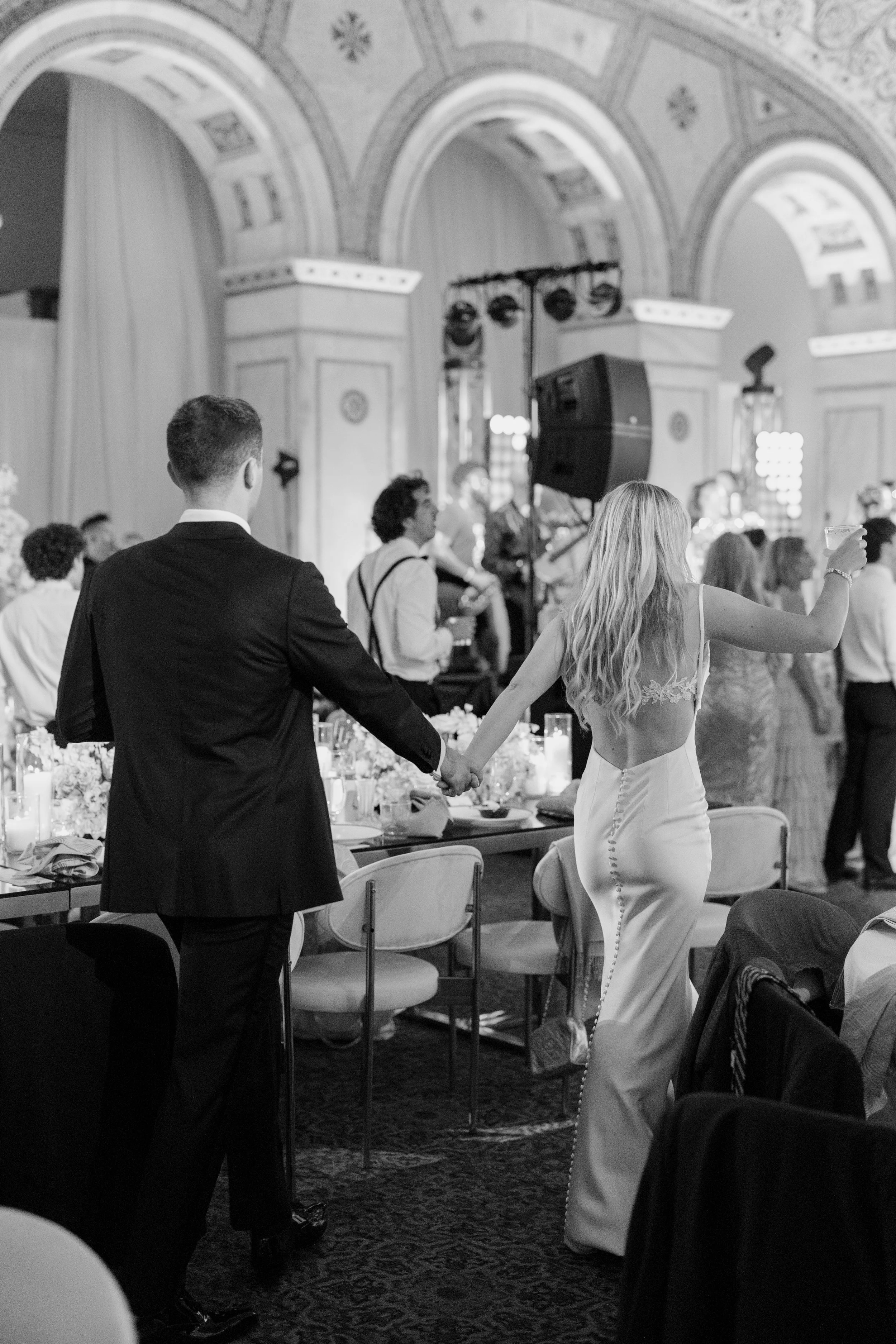 A man and woman holding hands at a formal event in a decorated hall, with other guests and musicians in the background.