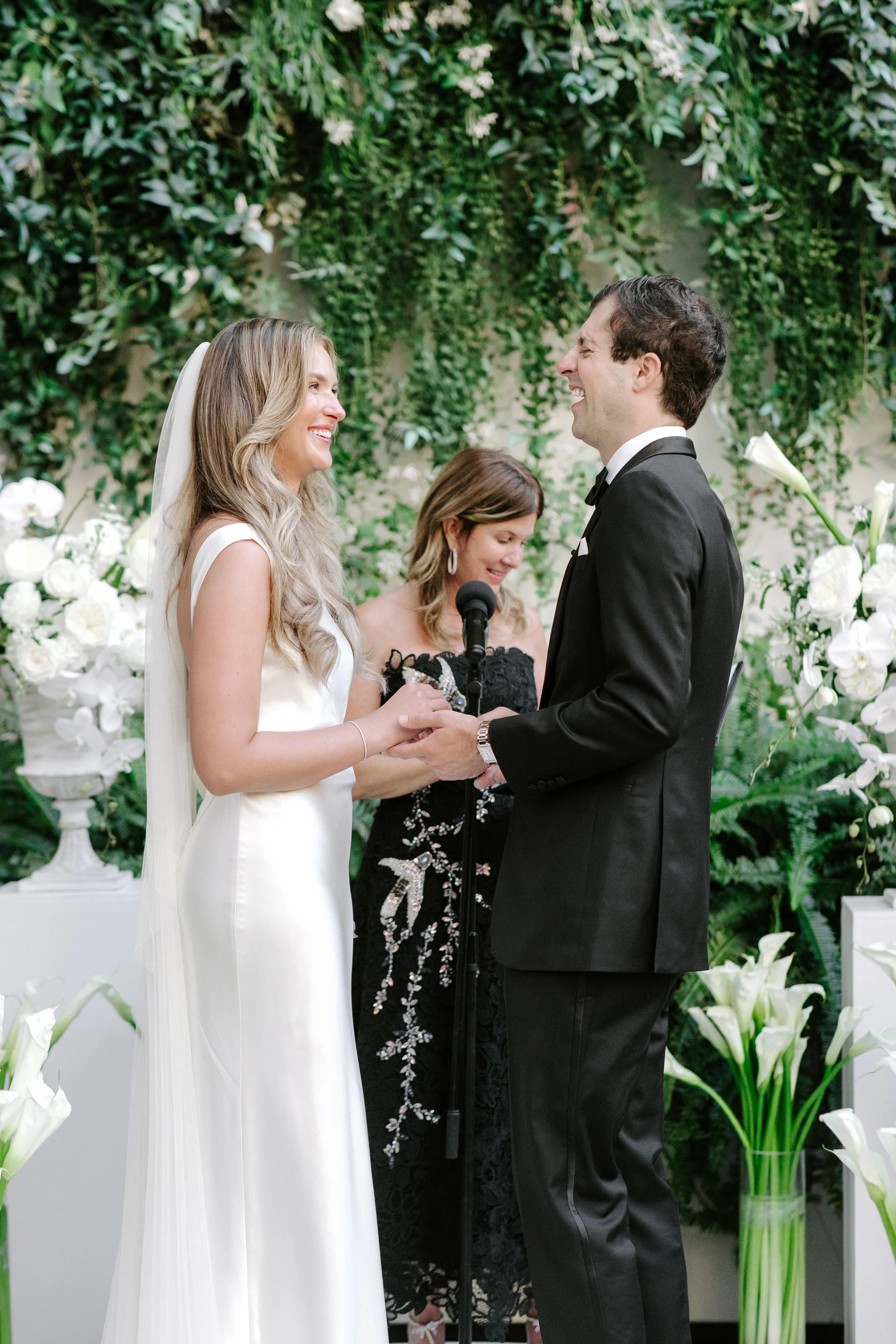 A bride and groom exchange vows during a wedding ceremony, standing face to face and smiling at each other, with a wedding officiant holding a microphone in the background surrounded by lush green foliage and white flowers.