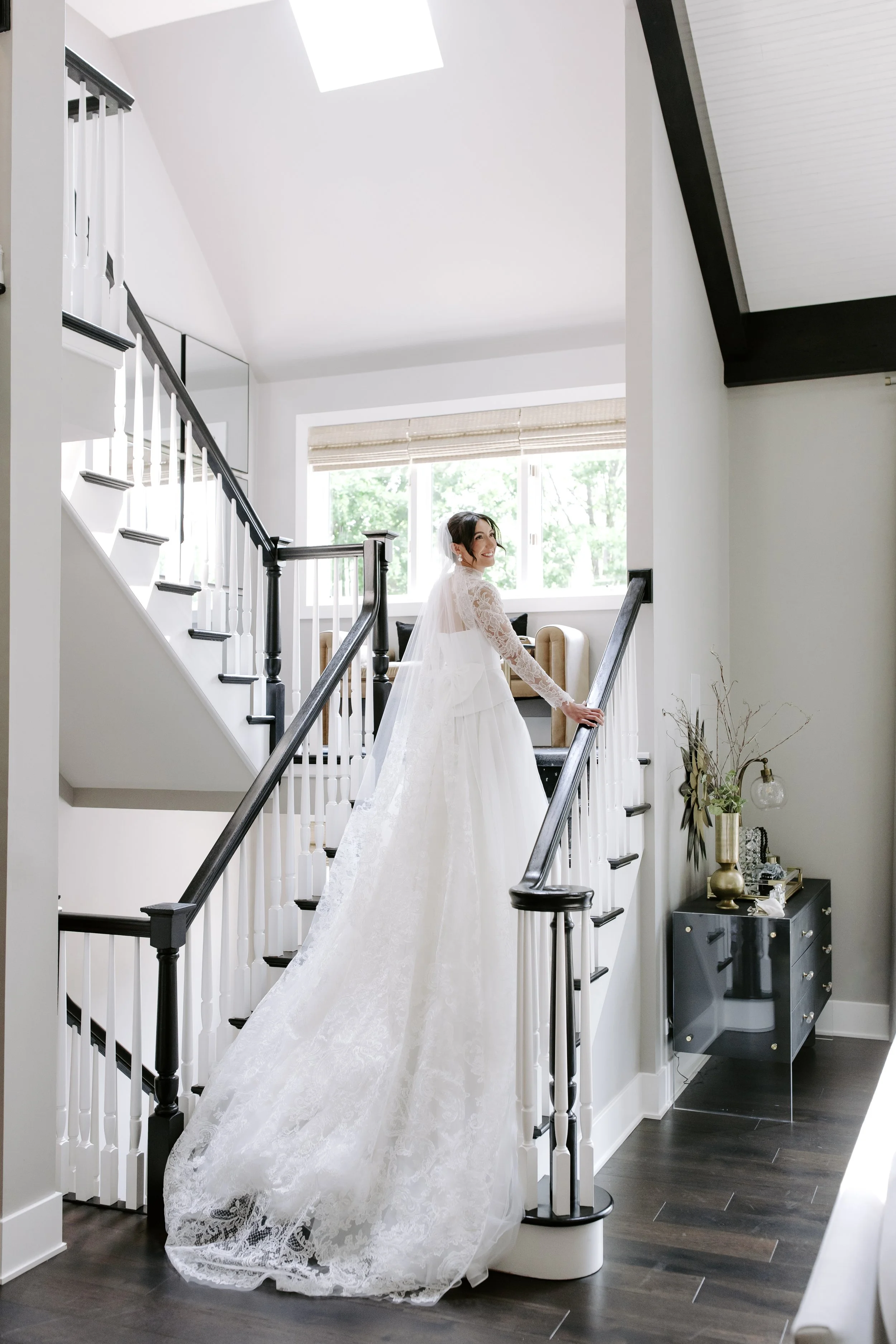 A bride in a white wedding dress and veil walking up a staircase indoors, smiling, with green trees visible through large windows in the background.