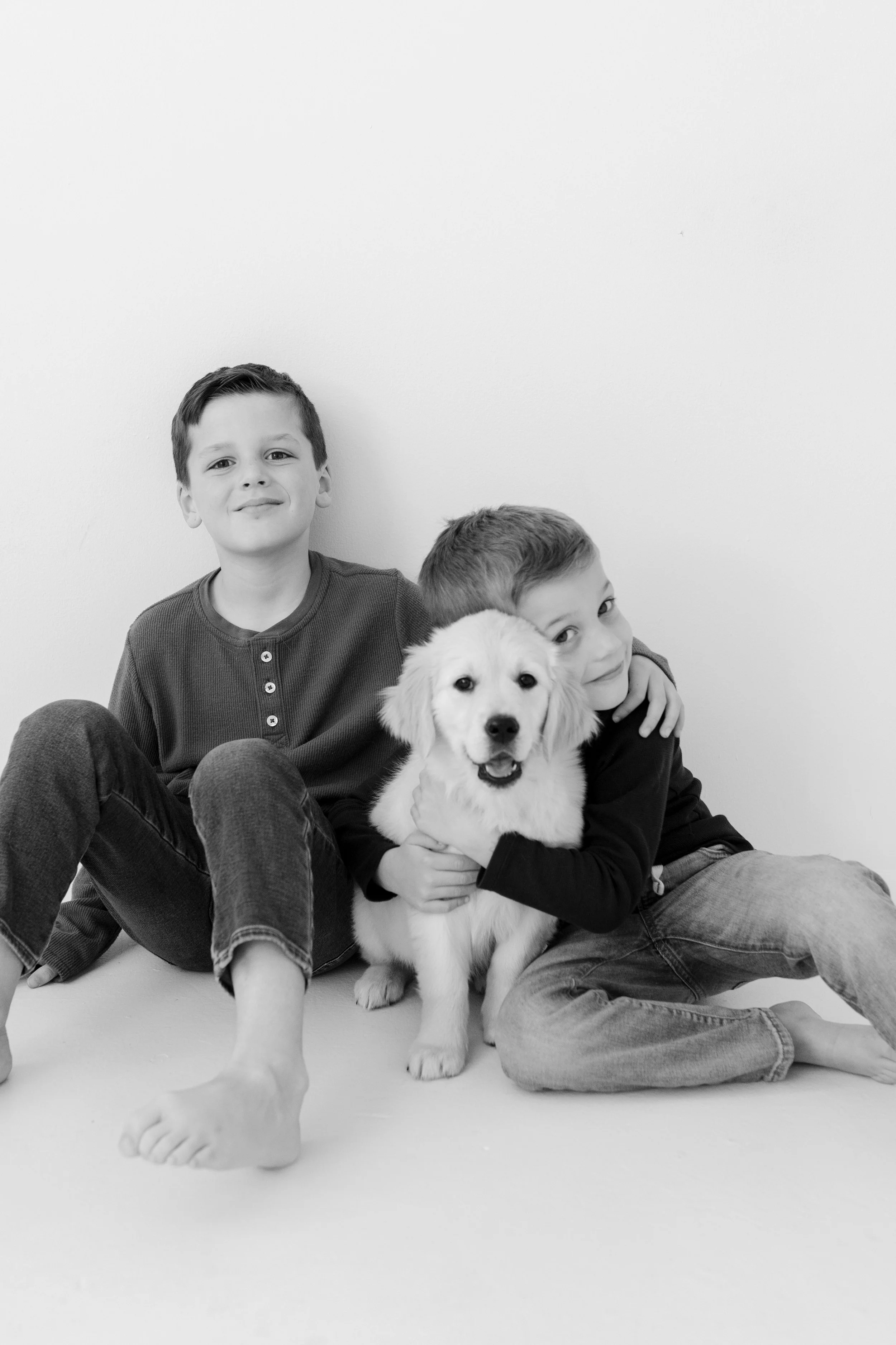 Two boys sitting on the floor with a golden retriever puppy, all smiling, in front of a plain white wall.