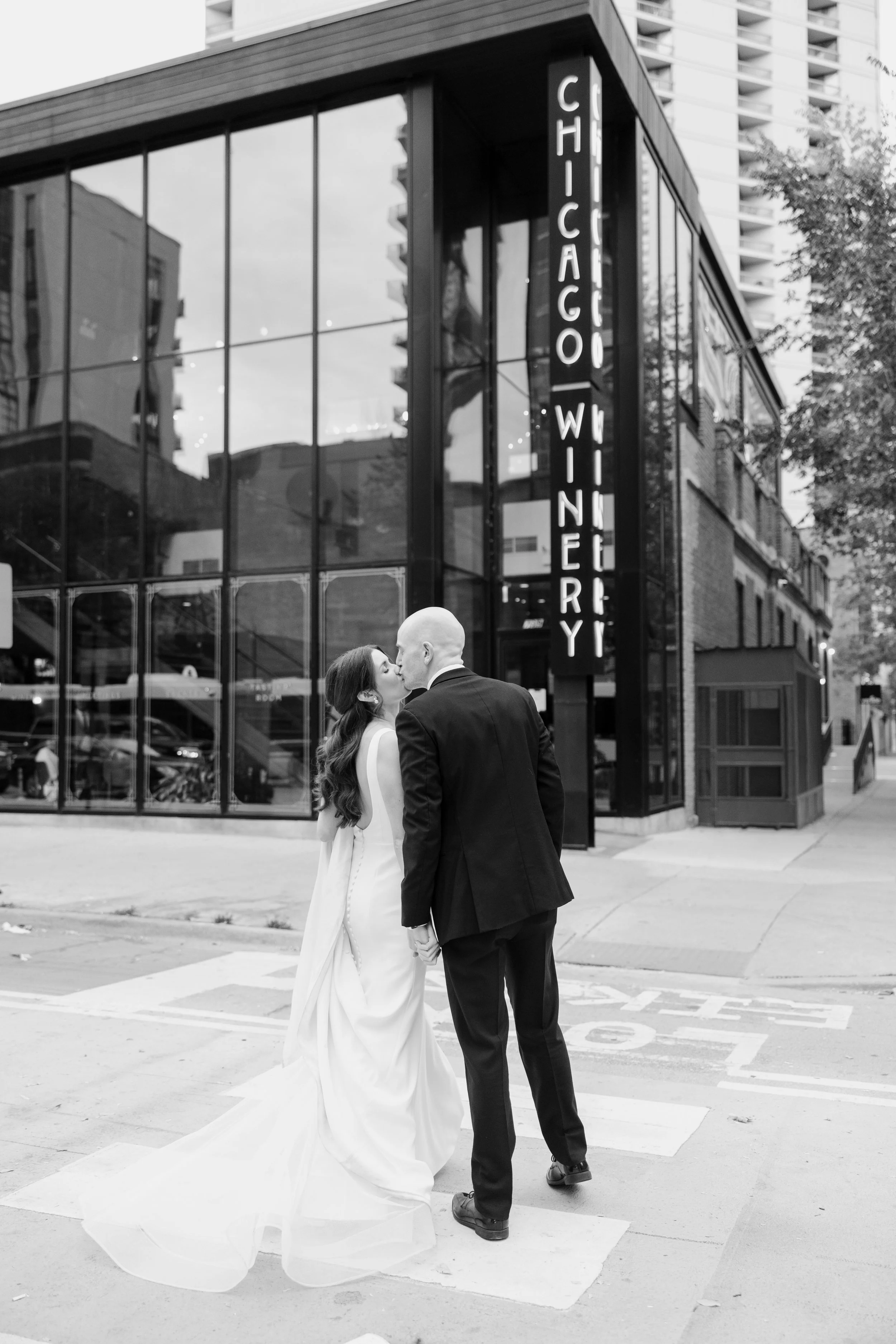 A bride and groom sharing a kiss on a city street in front of a neon sign that reads "Chicago Winery." The bride is wearing a long gown and the groom is dressed in a suit. The scene is captured in black and white.