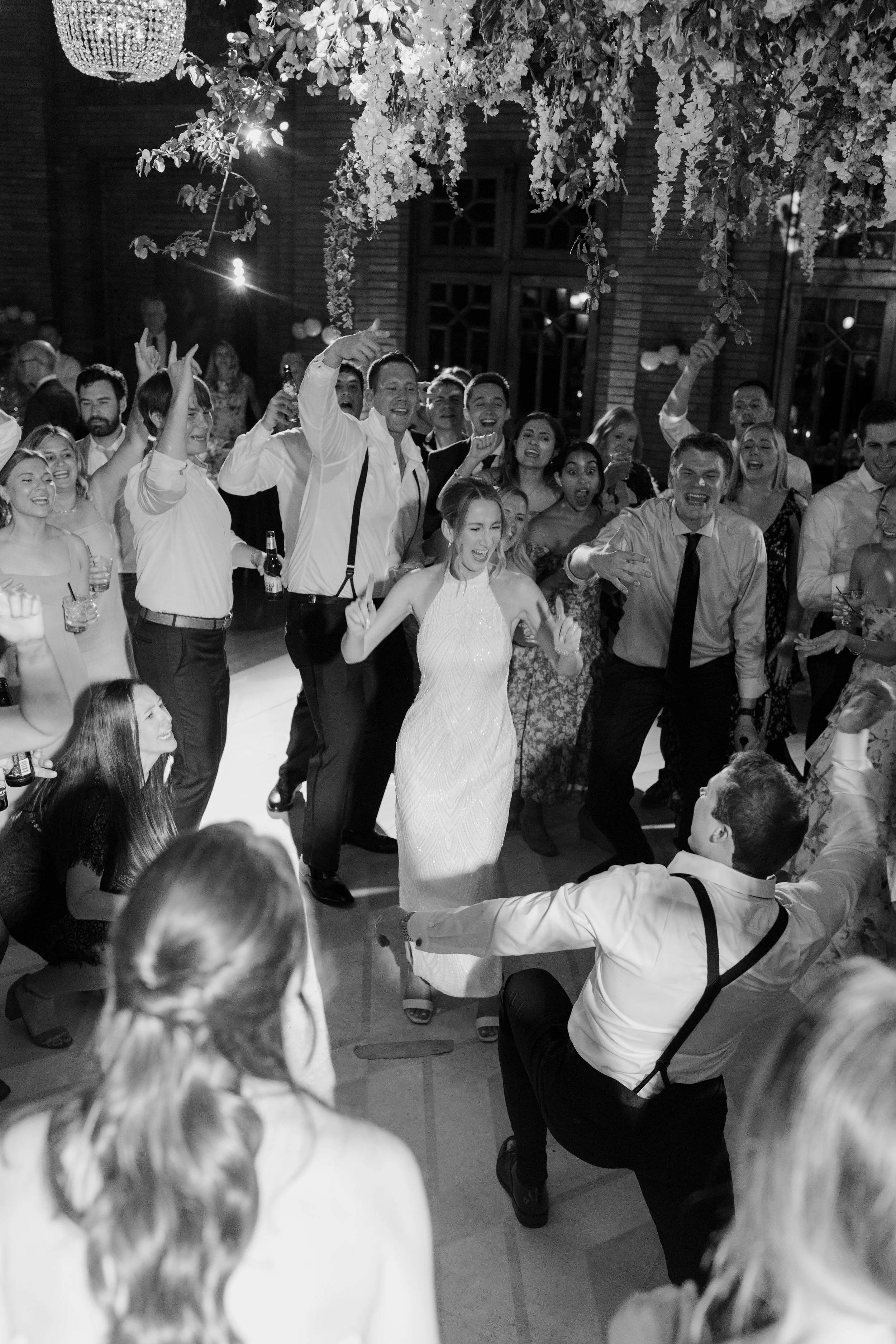 Black and white photograph of a lively wedding reception with guests dancing and celebrating. A woman in a white dress is the focal point, smiling and dancing. Decor includes hanging flowers and a chandelier.