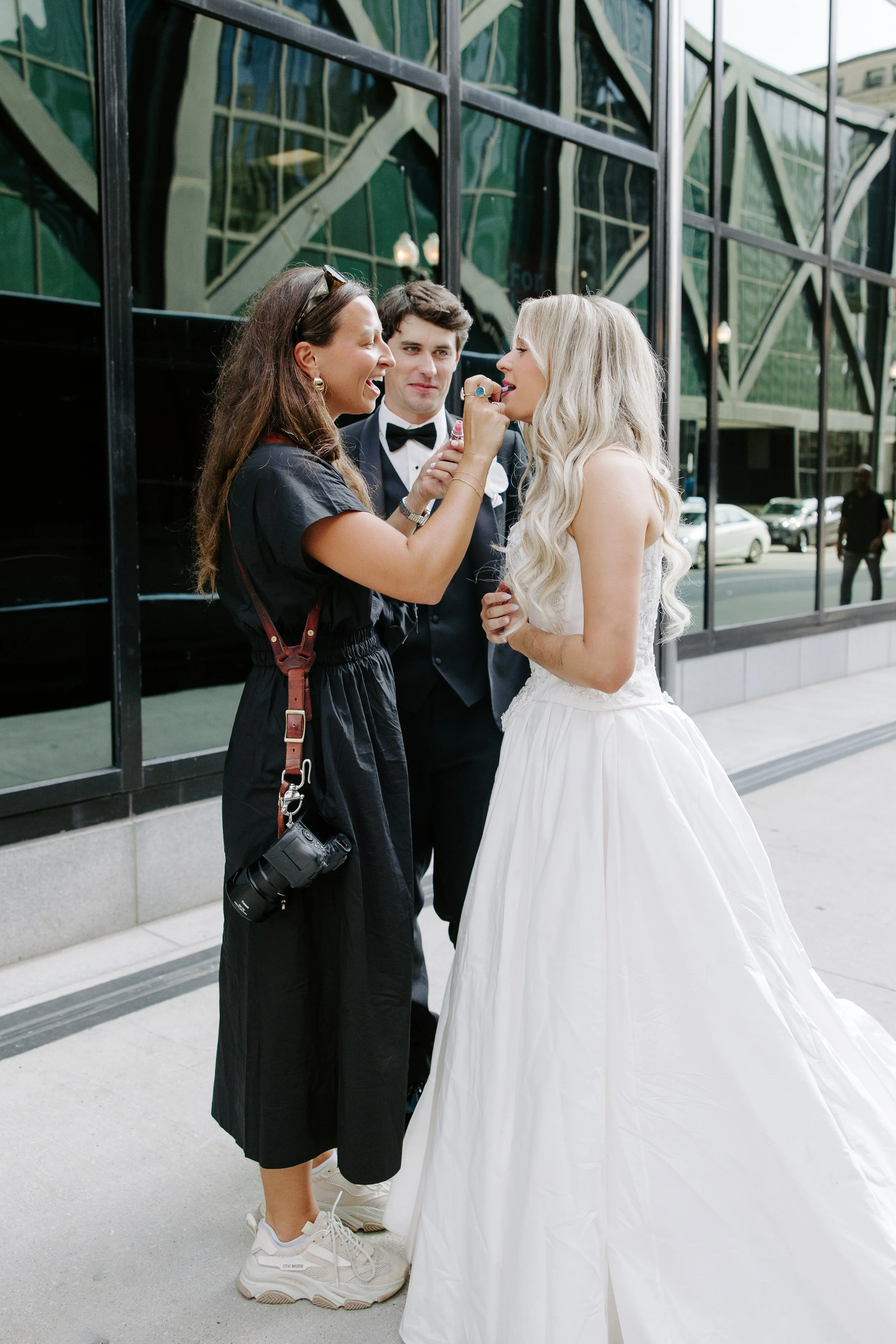 A woman applying makeup to a bride in a white wedding dress outside a modern glass building, with a man in a tuxedo watching.