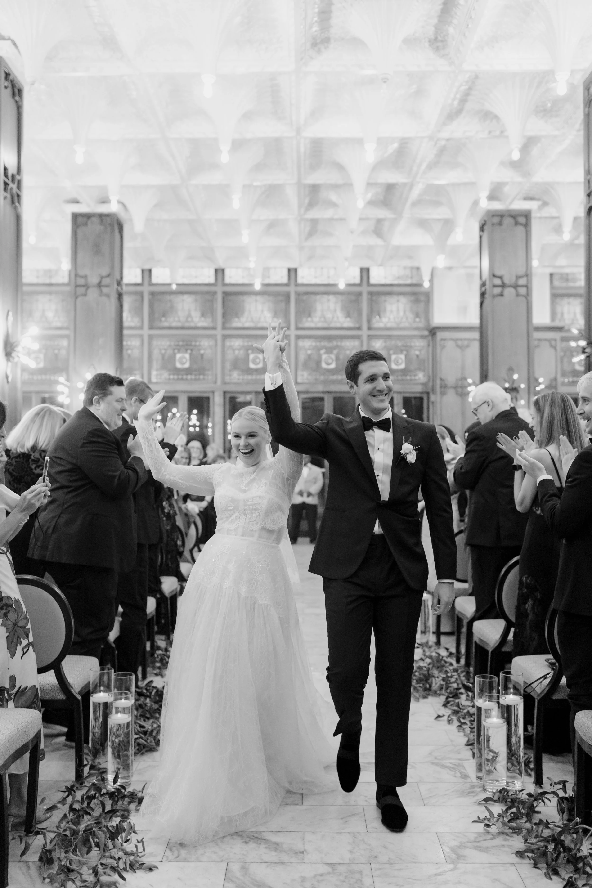 A bride and groom walking down the aisle at their wedding reception, smiling and holding hands, with the bride raising her hand in celebration. Guests are clapping and smiling on either side, in an elegant hall with ornate woodwork and decorative cei