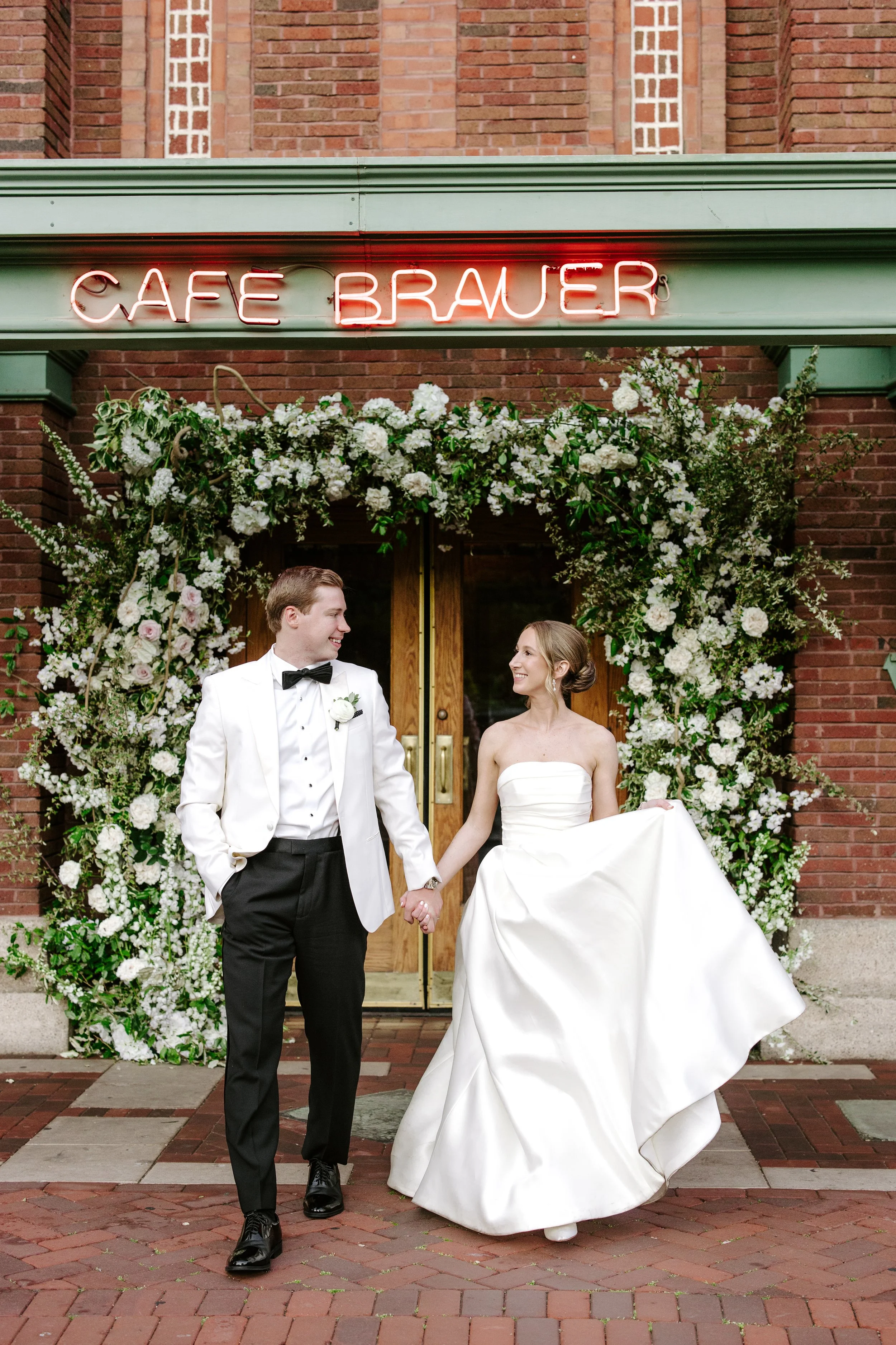 A bride and groom walking hand in hand outside a brick building, under a floral arch, with a neon sign that reads 'Café Brauer' above them.