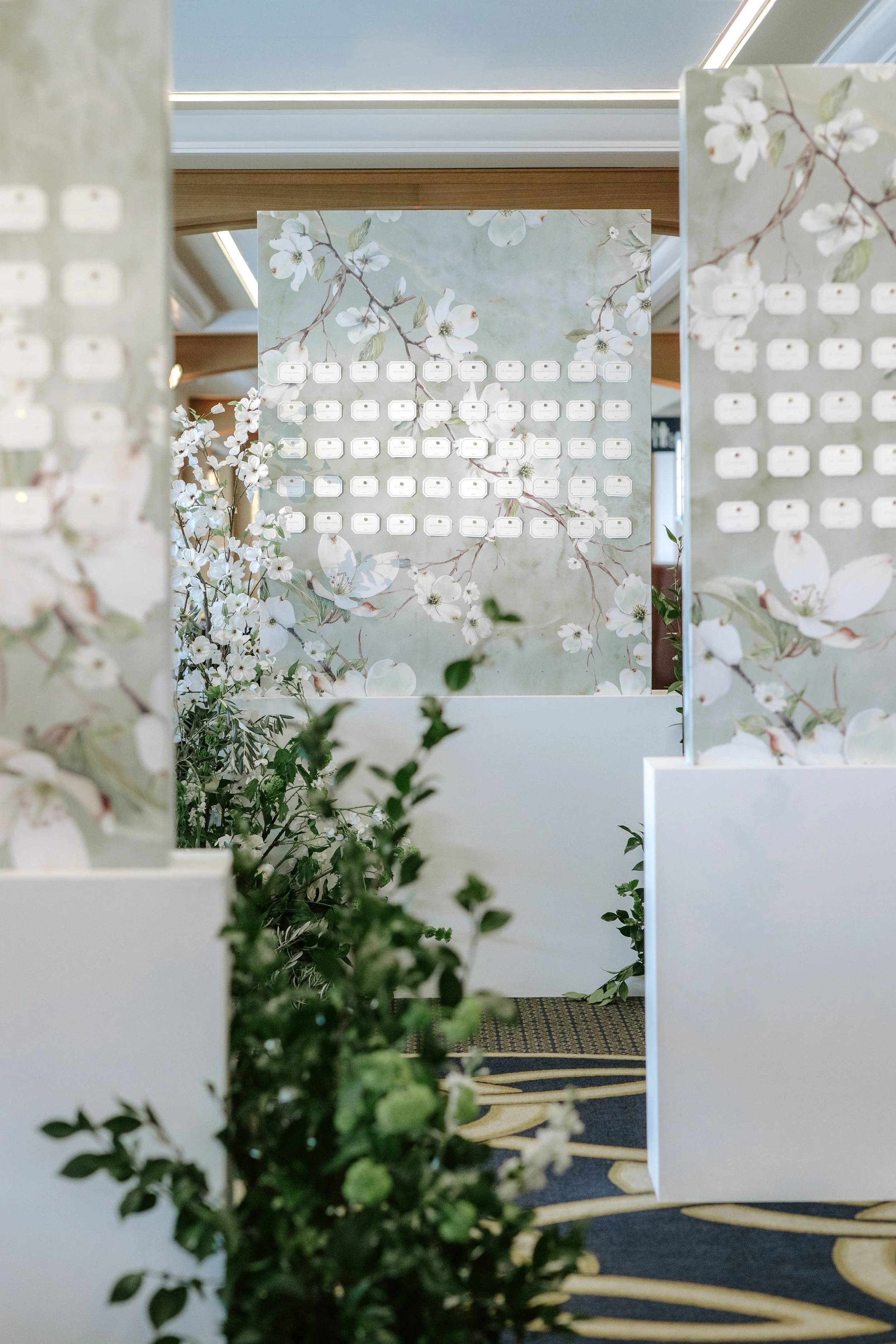 Decorative board with a floral design and small rectangular labels, surrounded by white flowers and green foliage, in an interior setting with a patterned carpet and ceiling window.