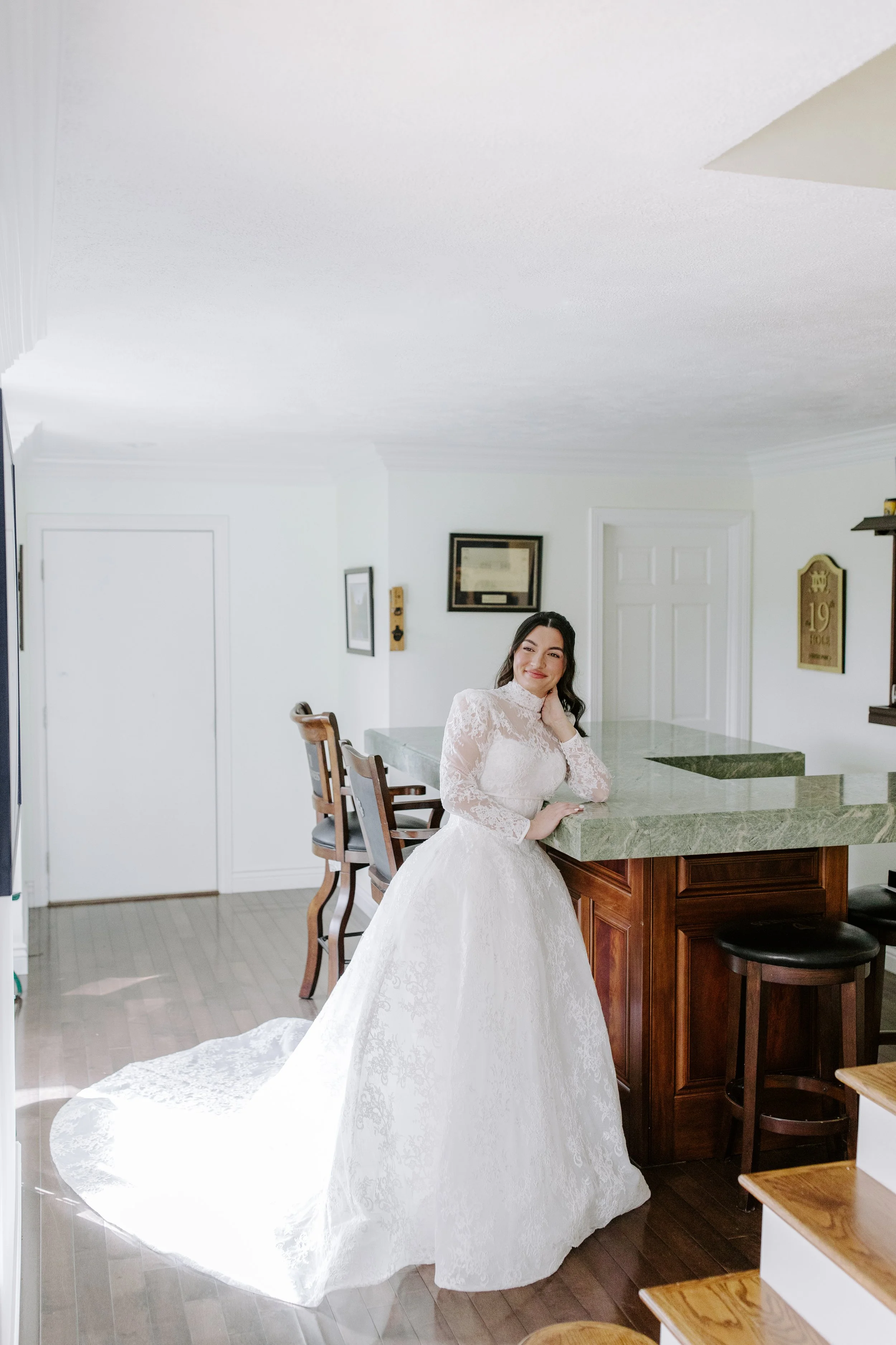 A woman in a wedding dress standing in a kitchen, smiling and leaning on a green marble kitchen island.