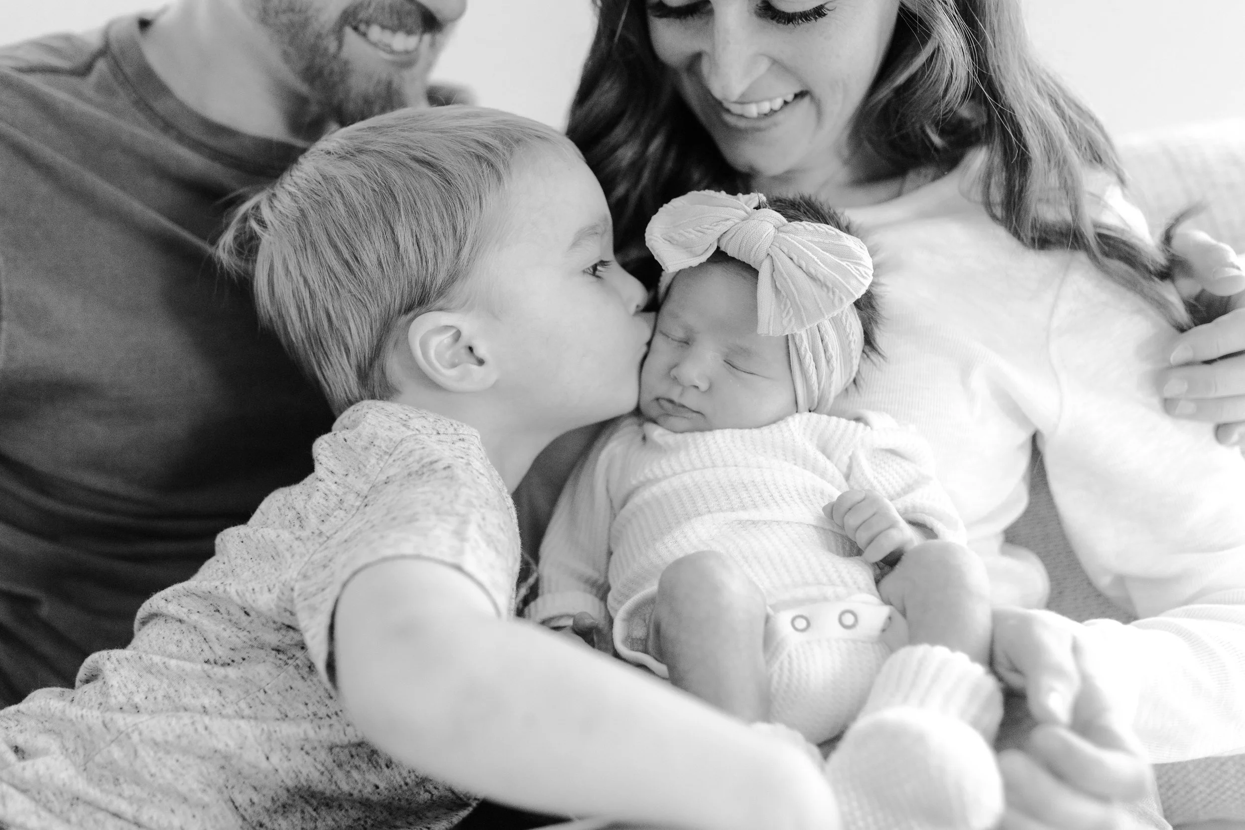 A family of four, including a father, mother, a young boy, and a newborn girl, showing affection as the father kisses the baby's forehead while the mother and boy look on smiling.