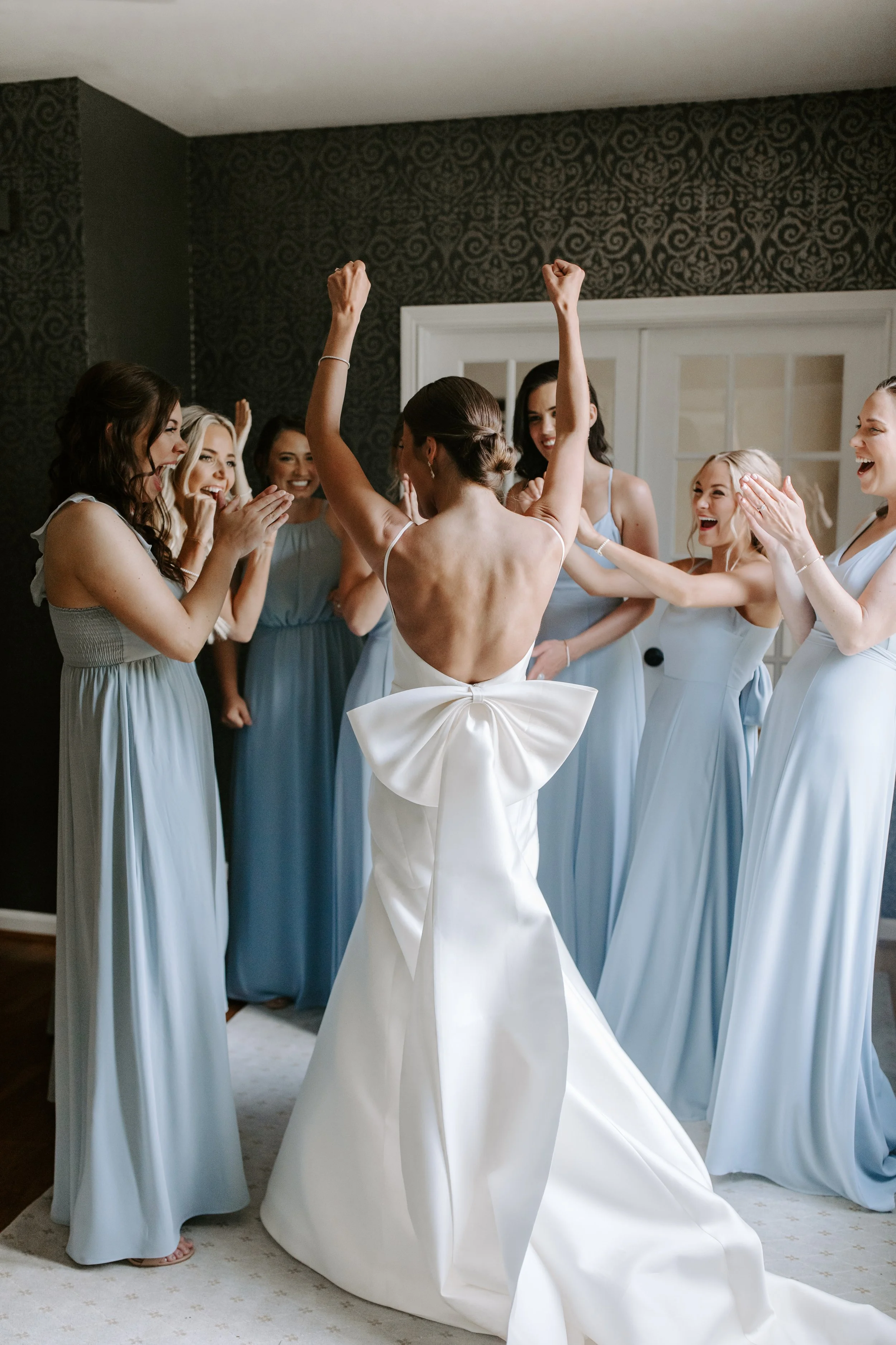 A bride in a white wedding dress with a large bow in the back celebrating with her bridesmaids in a room with dark patterned wallpaper.