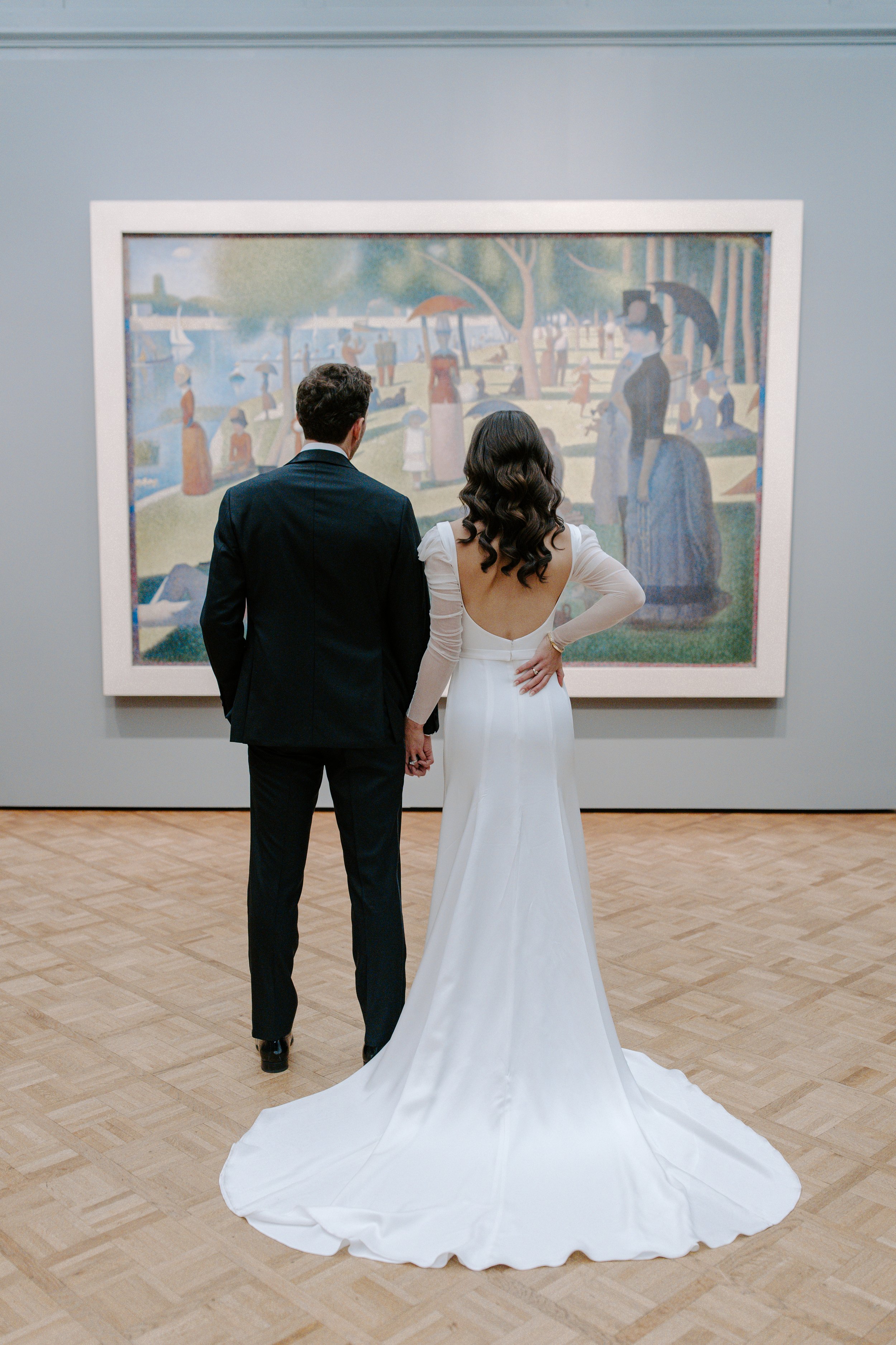 A couple dressed in wedding attire looking at artwork in an art gallery. The man is in a black suit, and the woman is in a white wedding dress with a long train.