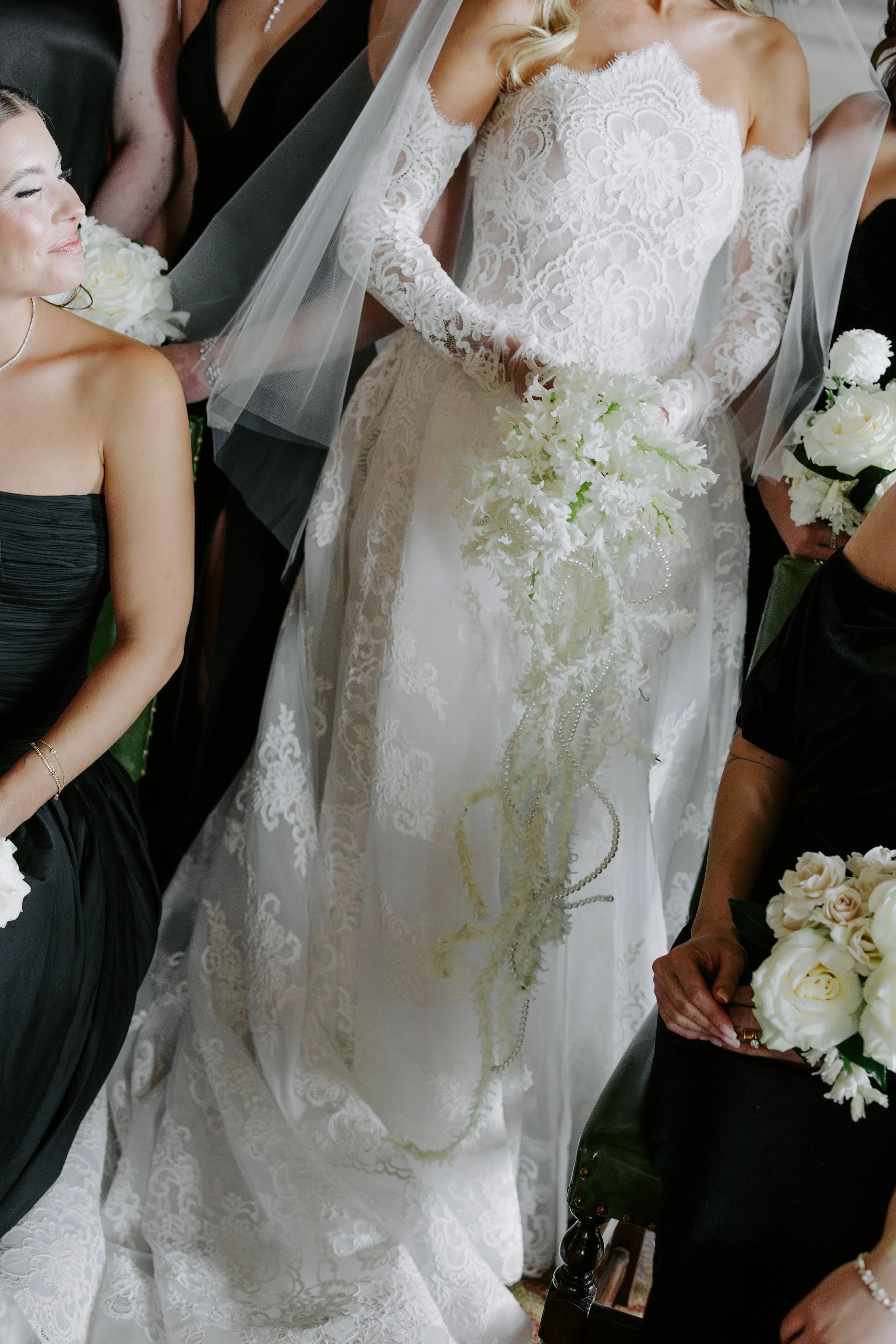 A bride in a white lace wedding gown holding a bouquet of white flowers, standing next to a woman in a black dress, among other women dressed in formal attire, during a wedding ceremony.