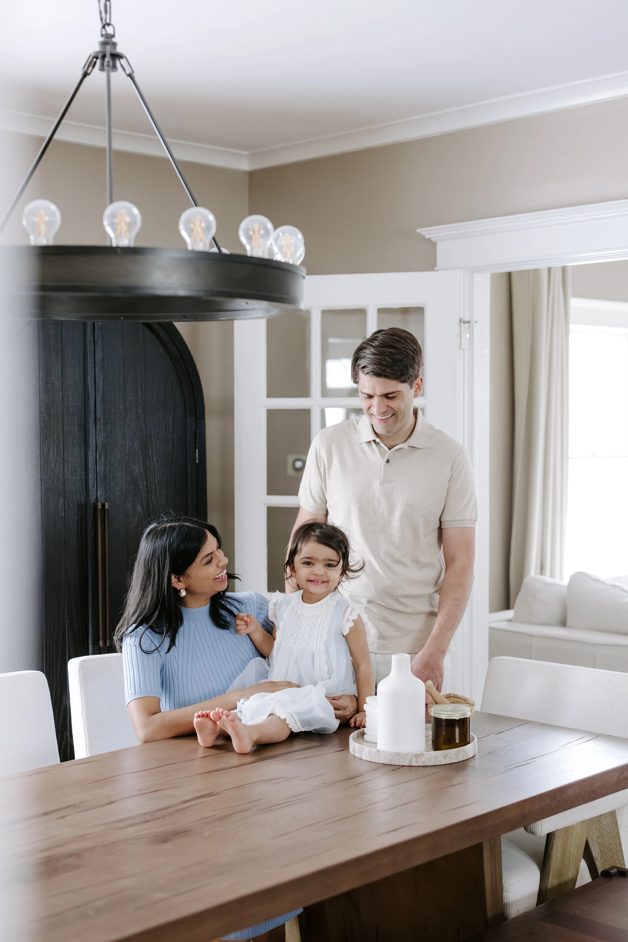 A family gathered around a dining table in a bright kitchen, with a woman holding a young girl and a man smiling nearby, on a sunny day.