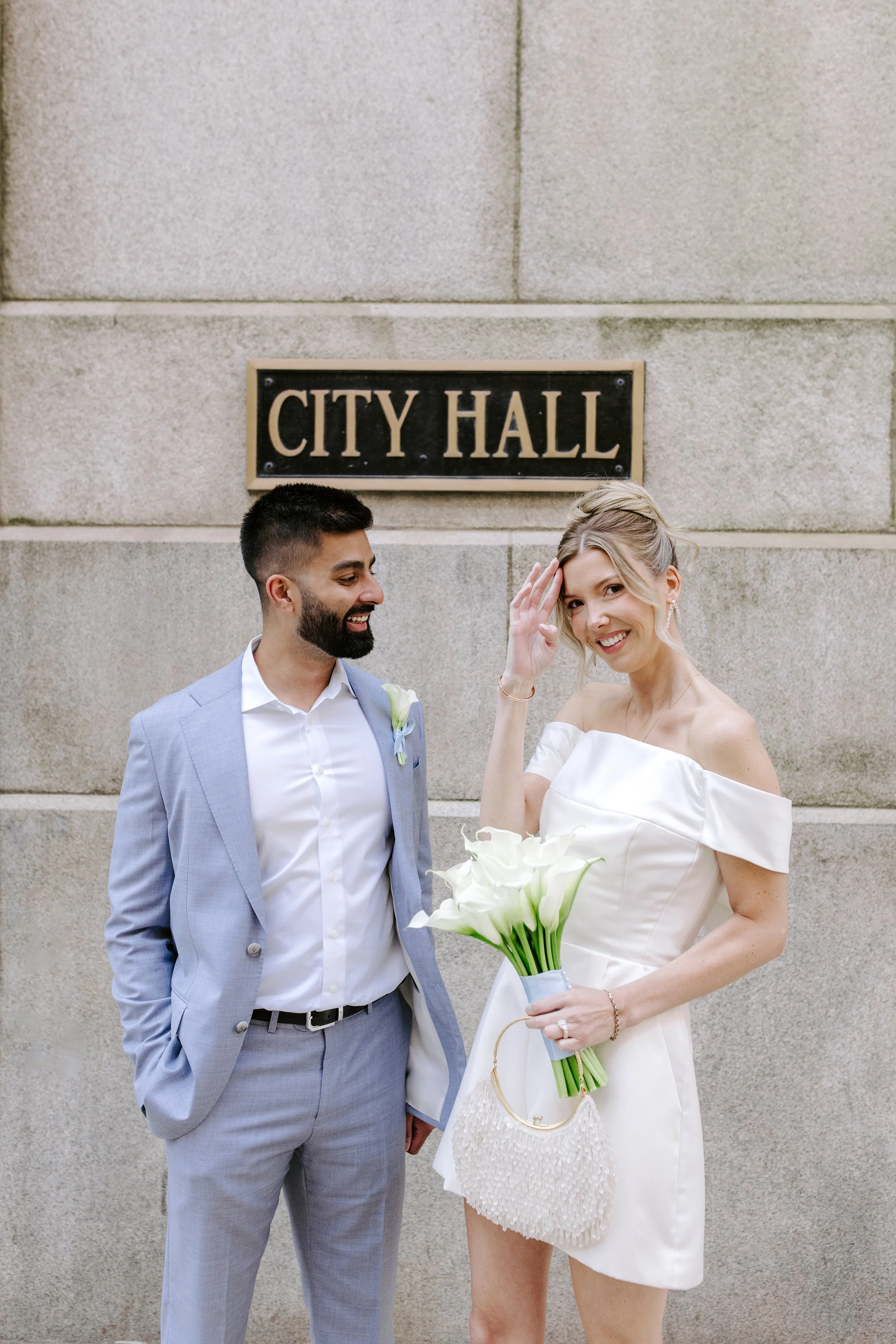 A man and woman in wedding attire standing in front of a stone wall with a sign that reads "City Hall." The woman is holding a bouquet of white calla lilies and is adjusting her hair, while the man is smiling and looking at her.