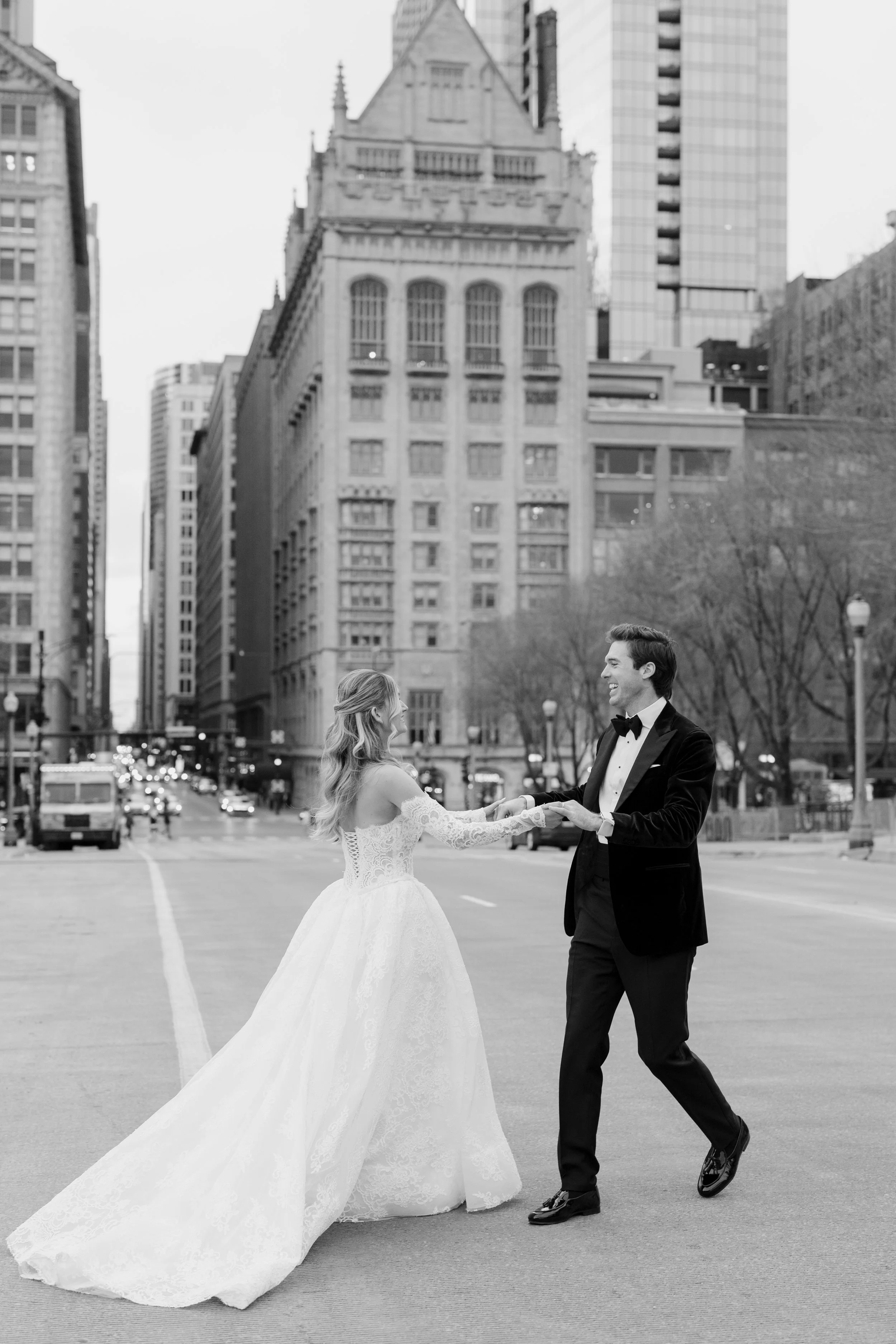 A bride and groom in formal wedding attire dancing in the middle of a city street, with tall buildings and leafless trees in the background.