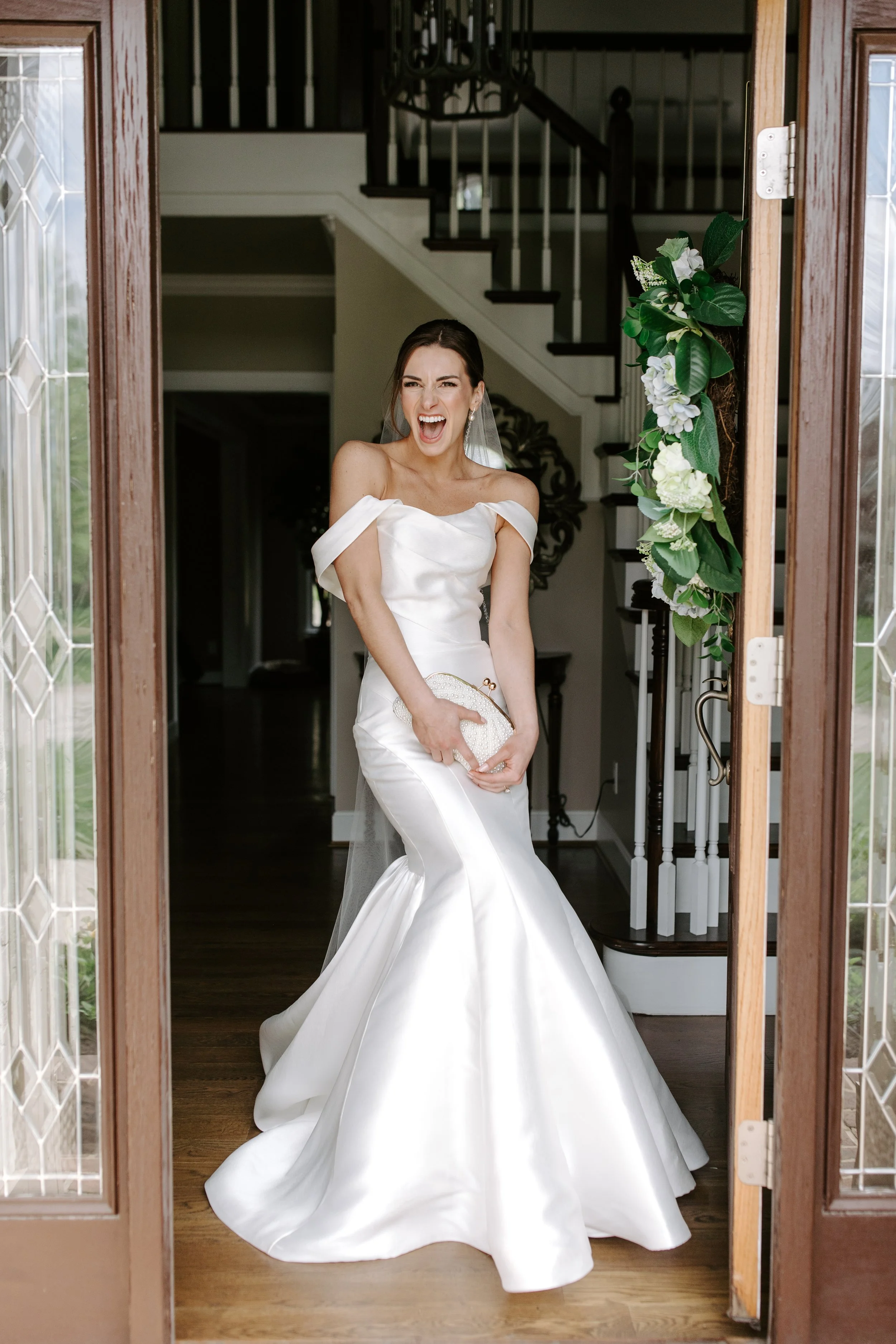 A woman in a white wedding dress standing in an entrance doorway, smiling and holding a clutch, with a staircase and decorative wall mirror in the background, and floral arrangements on the door frame.