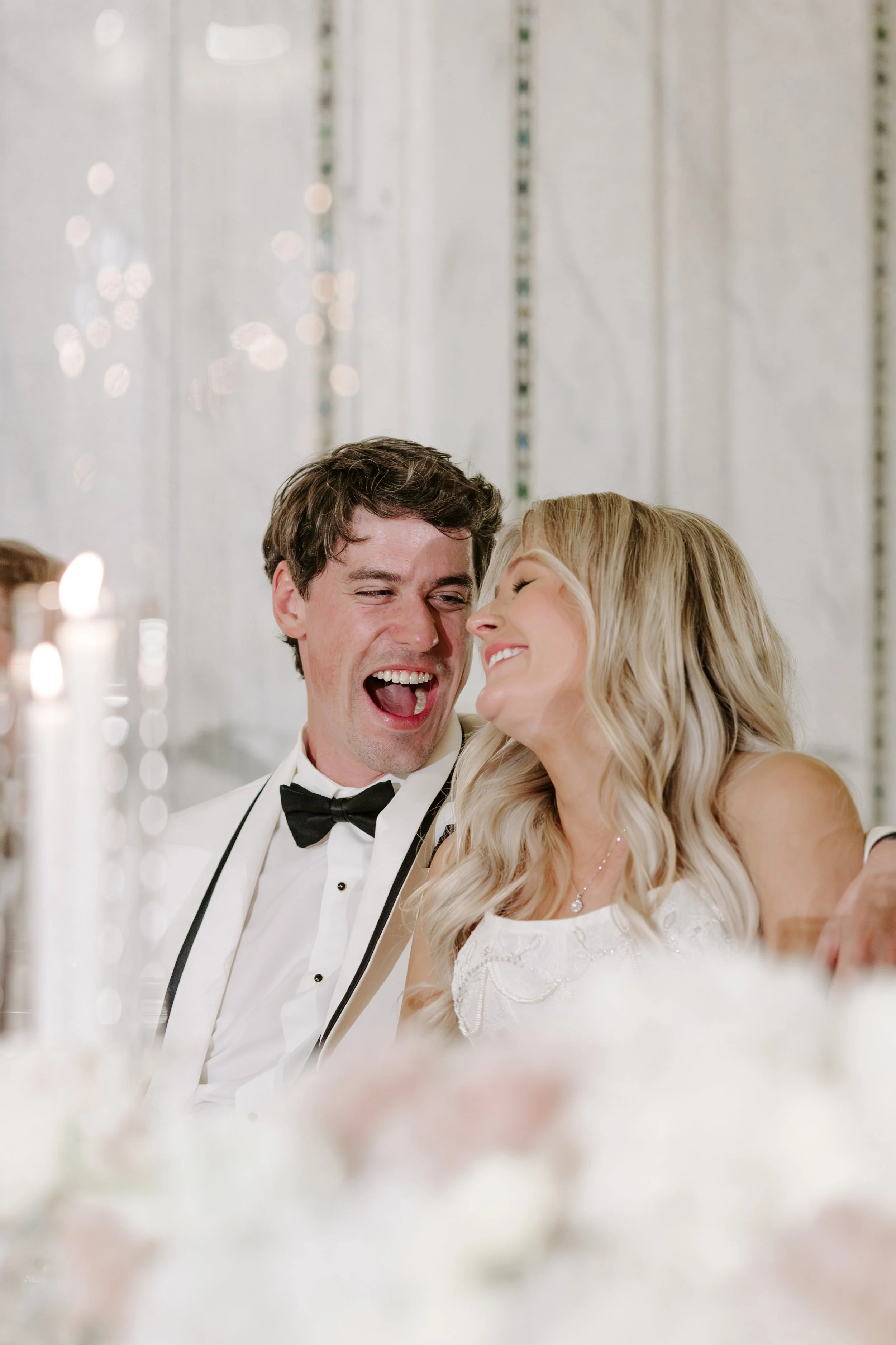 A couple dressed in formal wedding attire sharing a joyful moment, smiling, with blurred candles in the foreground and a white textured background.