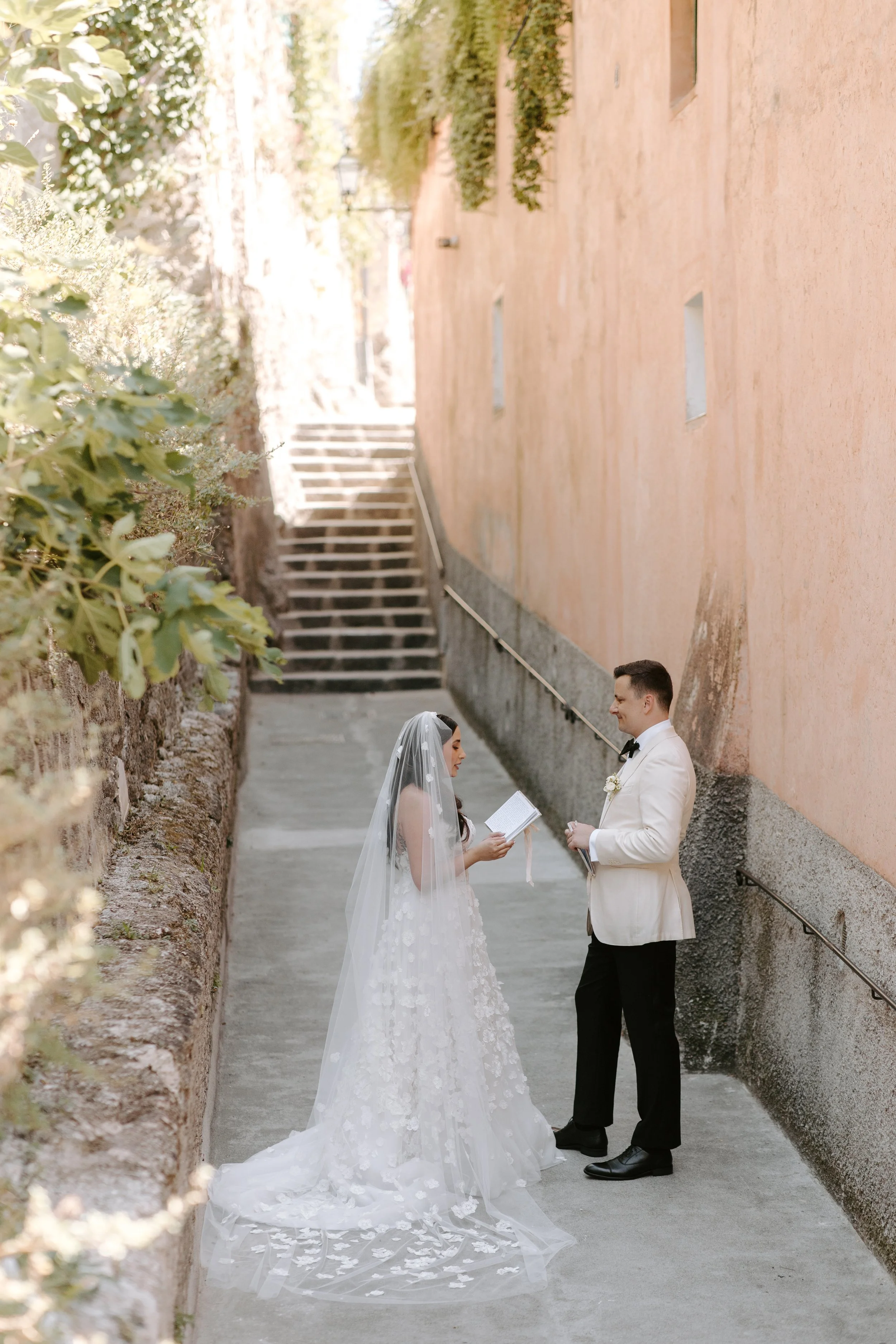 A bride and groom standing together outdoors during their wedding ceremony, facing each other with the bride holding a book and the groom smiling. The bride is wearing a white dress with lace details and a veil, and the groom is in a white tuxedo wit