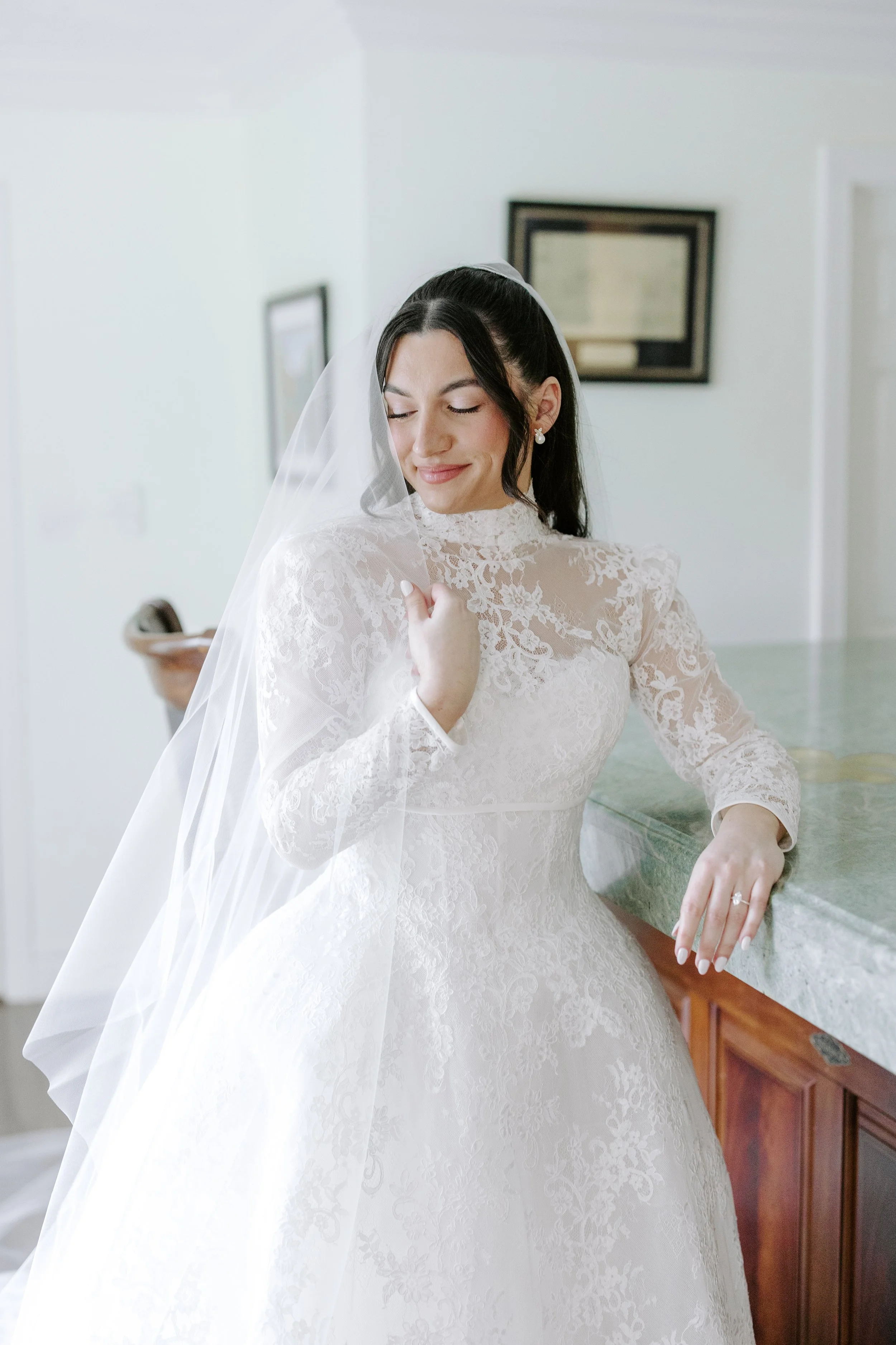 A bride in a white lace wedding dress with a veil, smiling softly, standing indoors near a green marble countertop with a wooden cabinet underneath, in a room with framed pictures on a white wall.