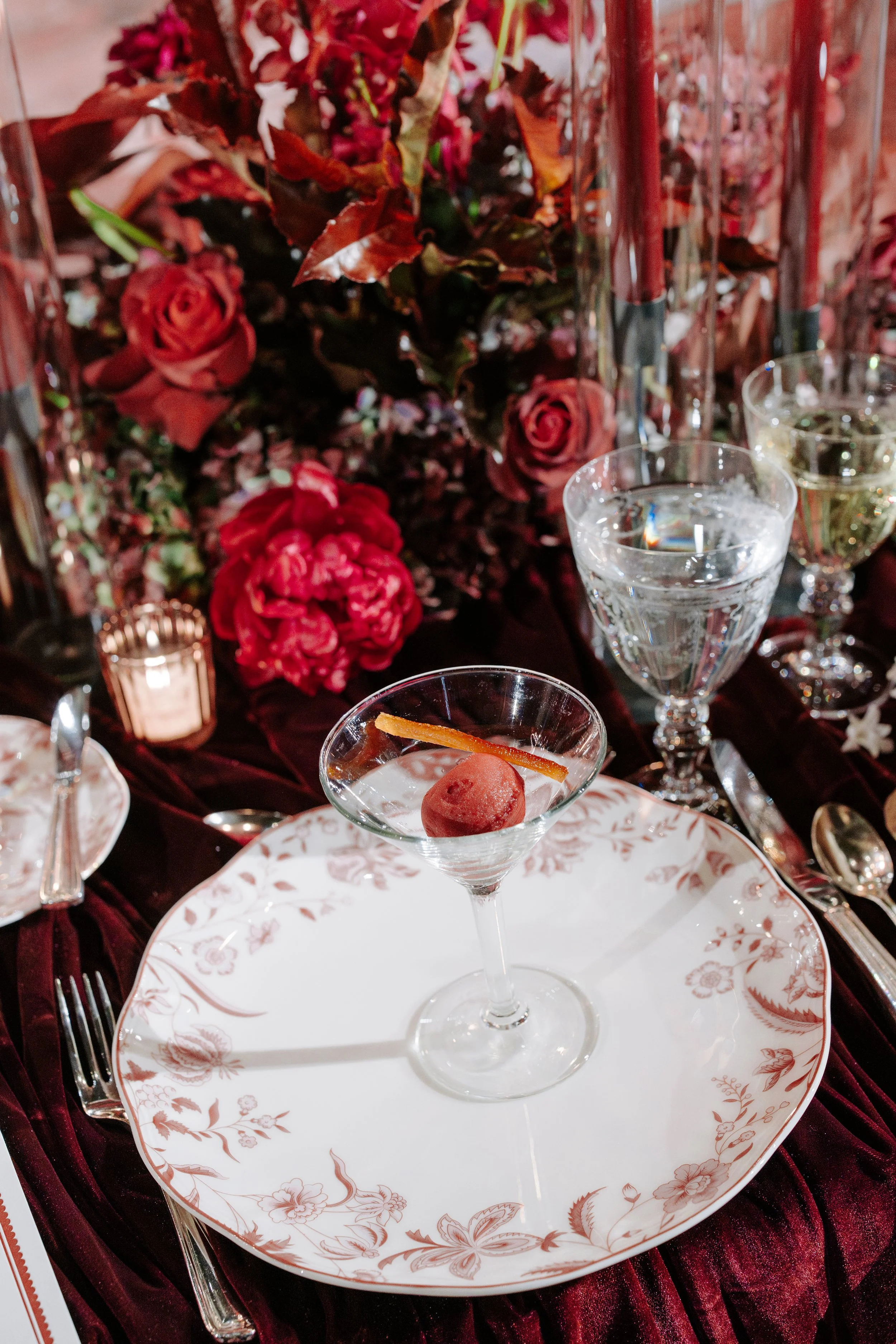 A formal dining setup with a floral arrangement background, featuring a glass of water, a glass of wine, and a martini glass with a cocktail and garnish on a decorative plate.
