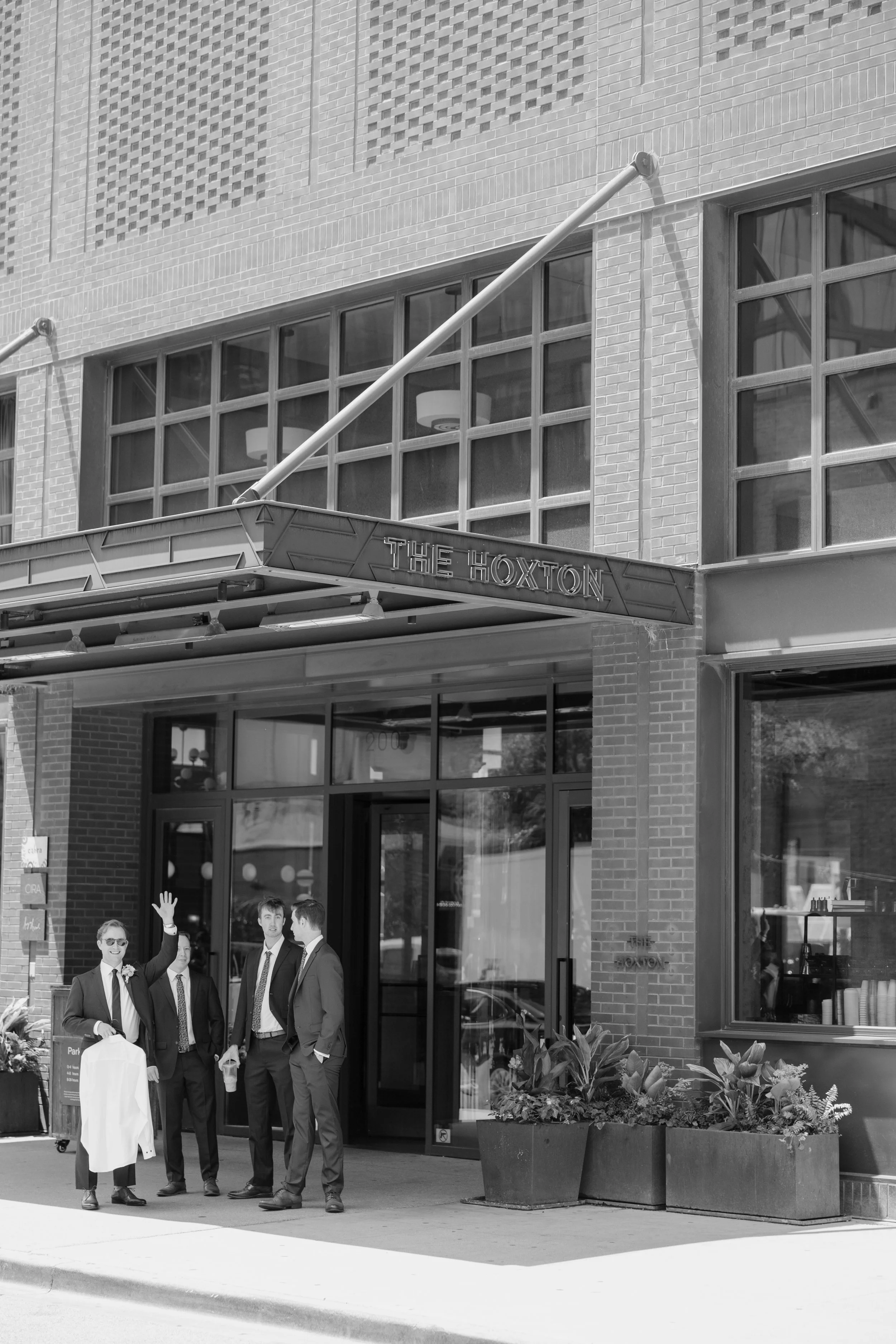 People dressed in formal attire standing and talking outside a building called 'The Hoxton' with large windows and a decorative brick exterior, and a large planter with flowers in front of the storefront.