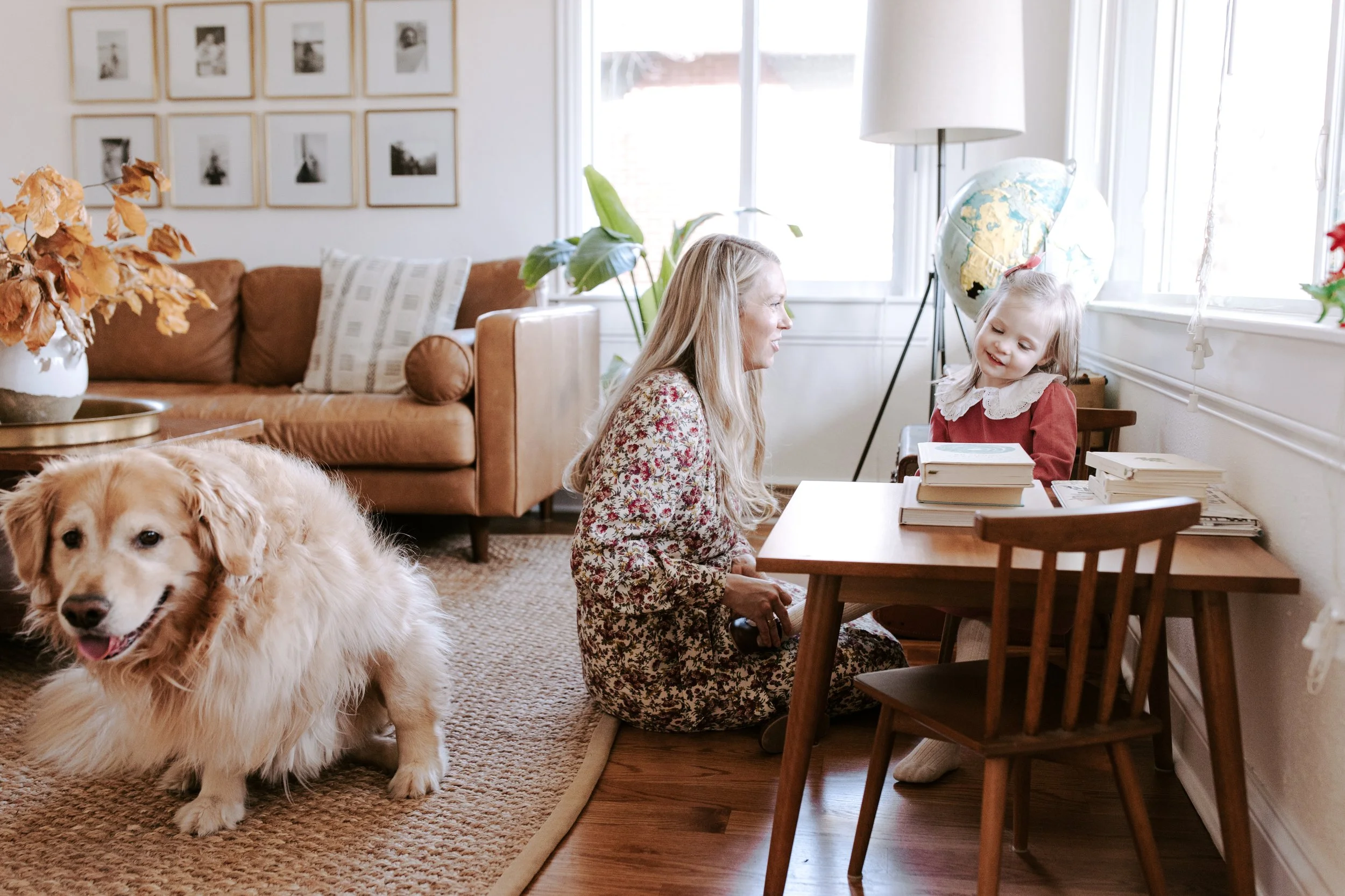 A woman and a young girl sitting at a wooden dining table, with a large dog nearby. The room is well-lit with natural light, has a bookshelf, a globe, a large plant, and framed photos on the wall.