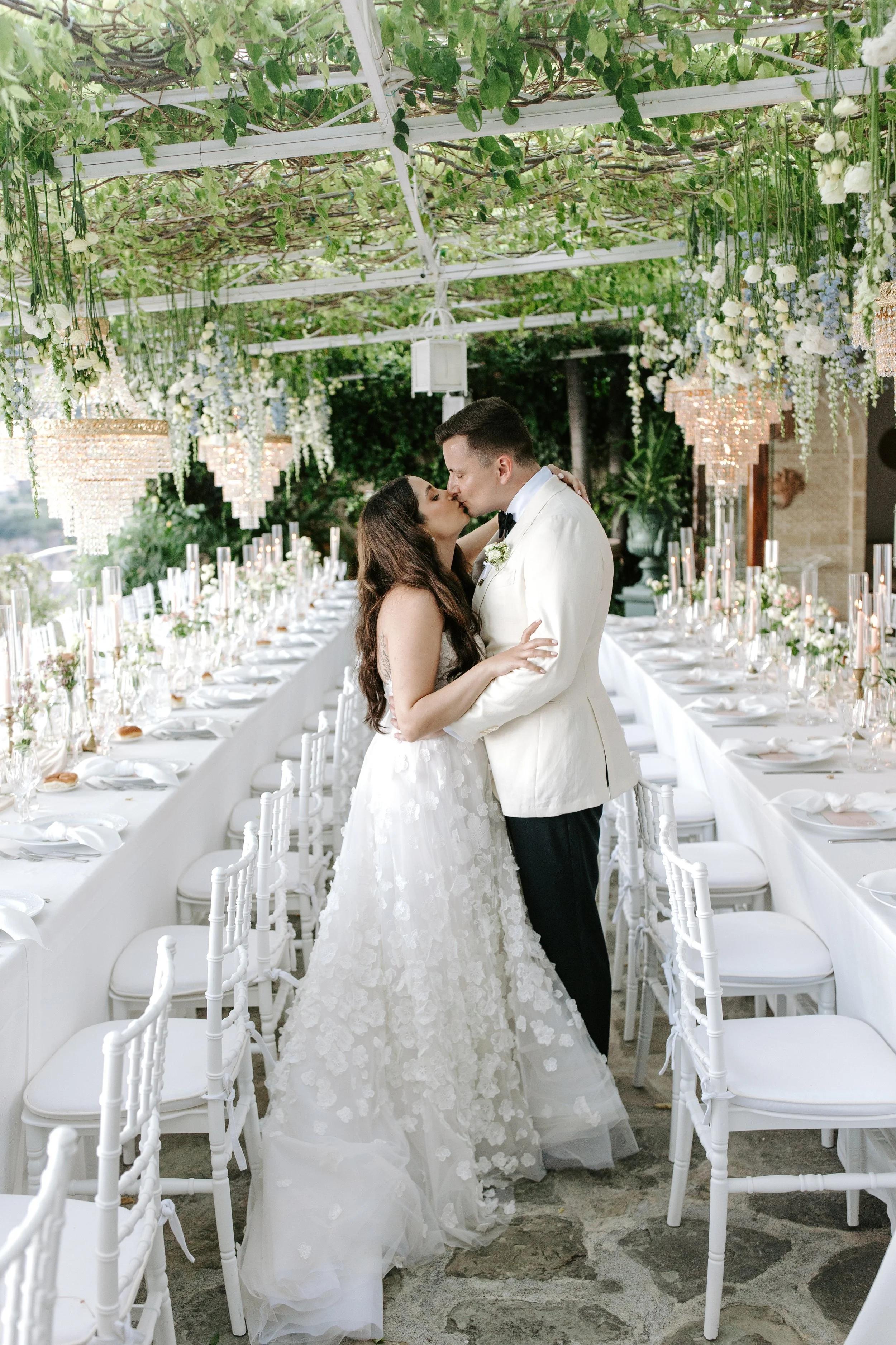 Bride and groom sharing a kiss at their wedding reception with long tables decorated with white flowers and candles, under a lush green floral canopy.