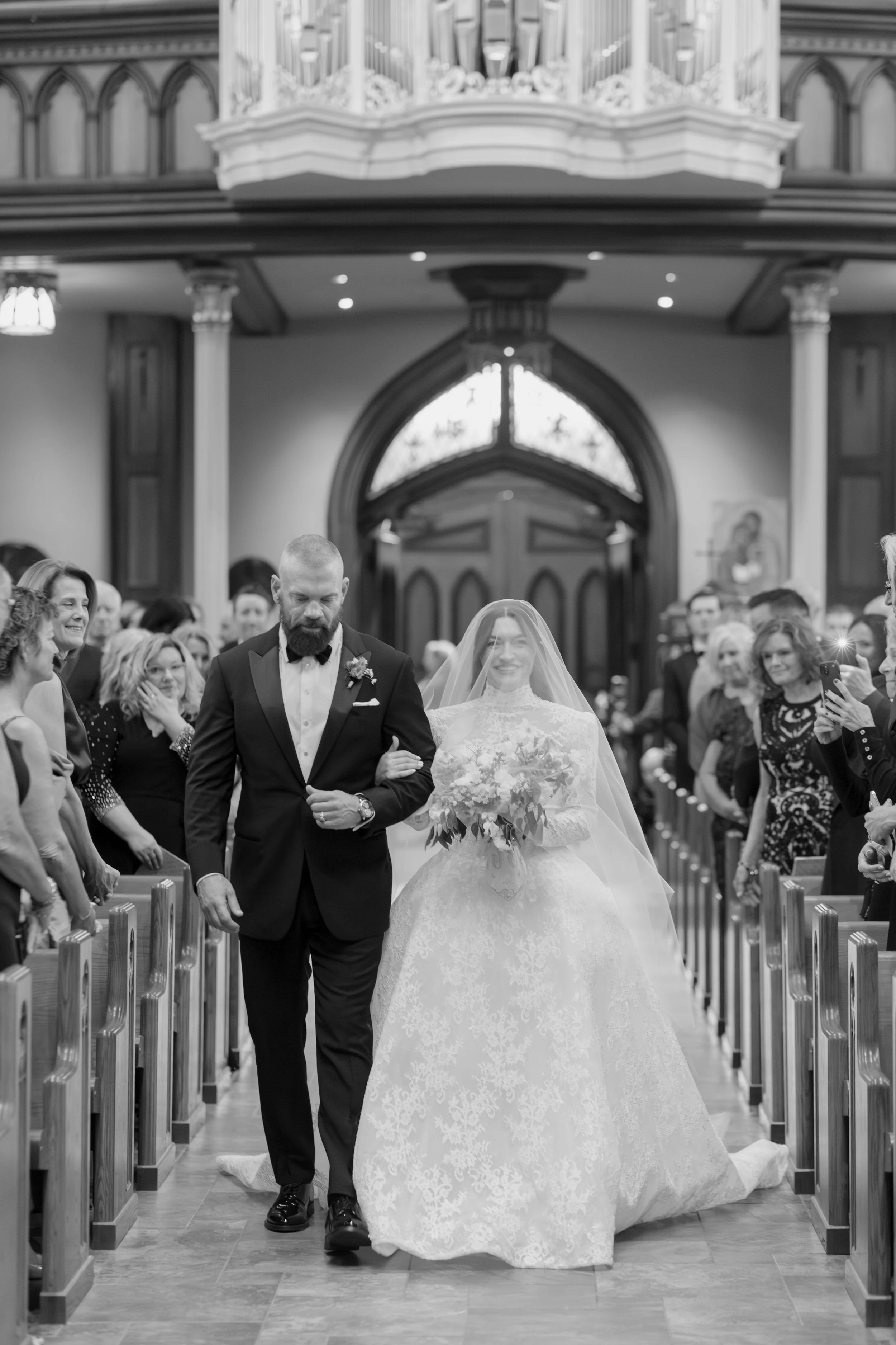 A bride and groom walking down the aisle in a church, surrounded by guests. The bride is wearing a lace wedding gown and veil, holding a bouquet of flowers. The groom is dressed in a black tuxedo with a bowtie and boutonniere. Guests are watching and