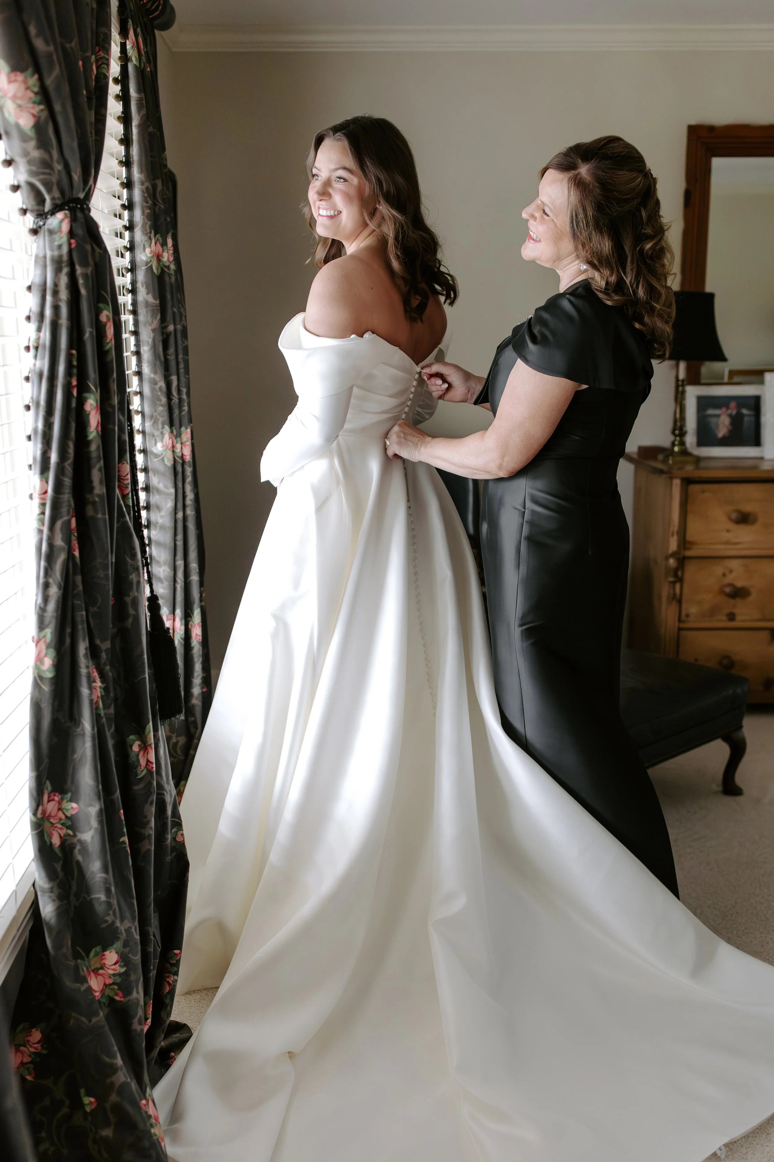 A woman in a white wedding dress is smiling as another woman buttoned up her dress. They are standing near a window with floral curtains in a cozy room.