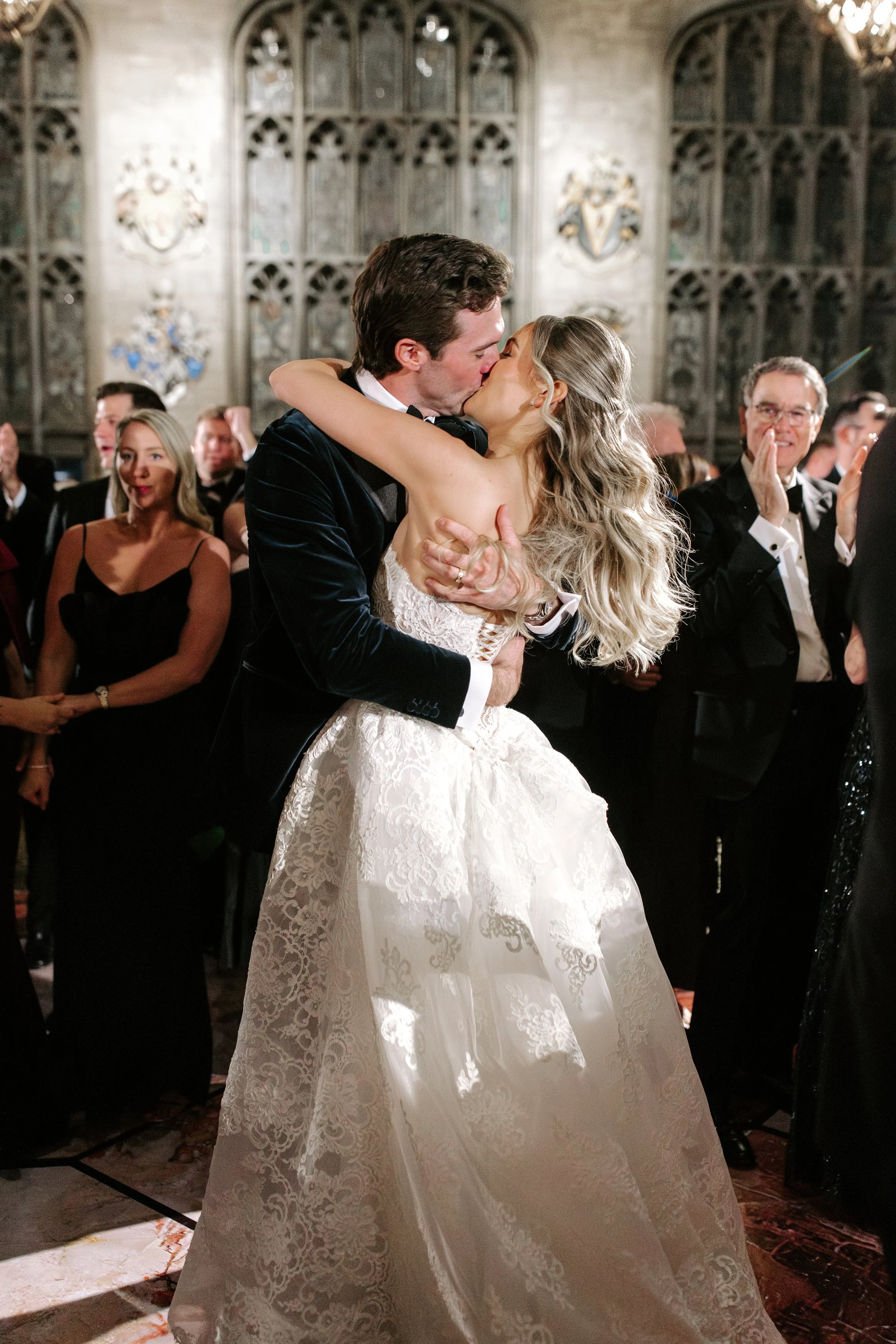 A bride and groom kissing during their wedding ceremony in a historic hall with stained glass windows. The bride wears a lace wedding gown with long, wavy blonde hair. The groom is in a dark tuxedo. Guests are standing and clapping in the background.