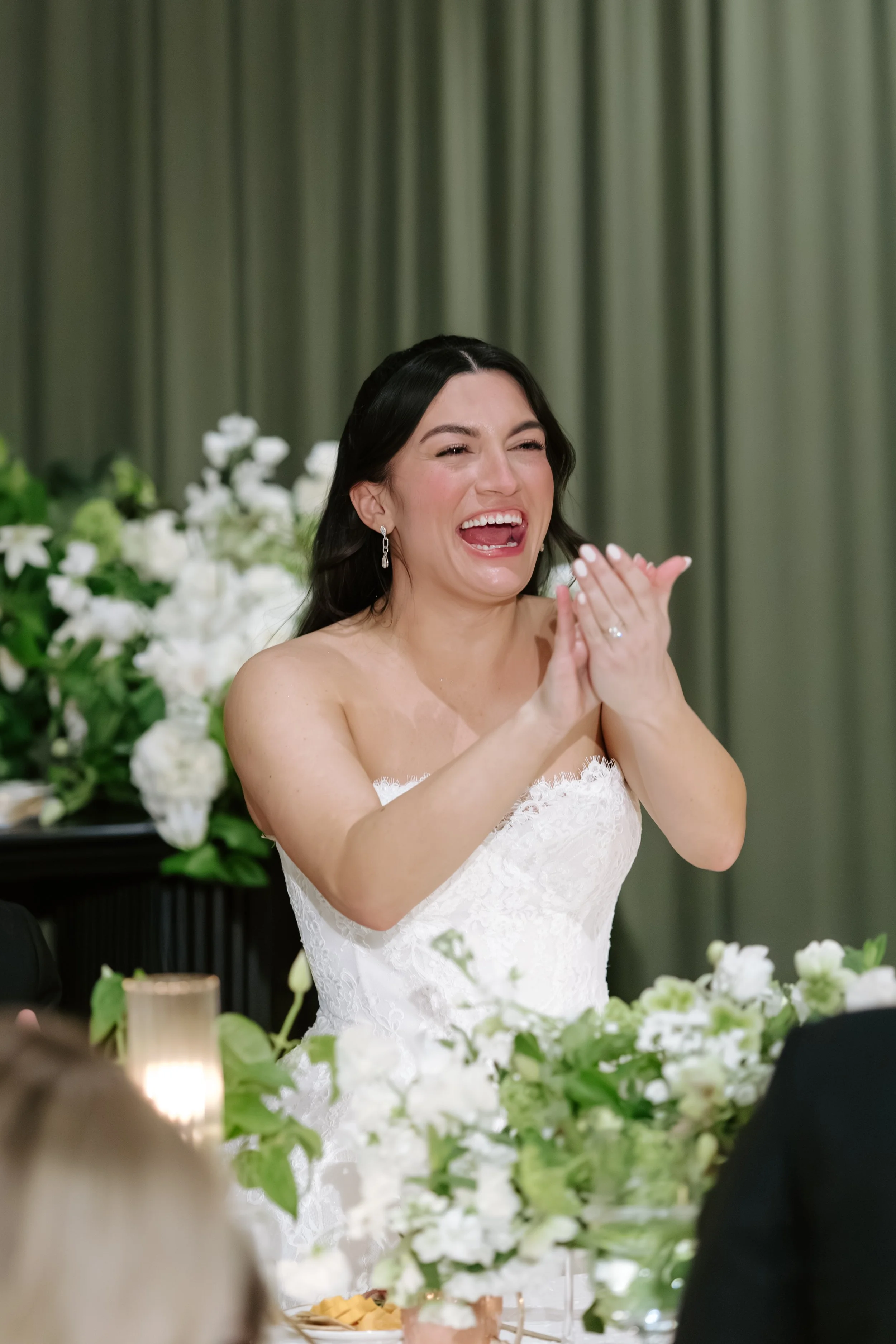 A woman in a white wedding dress is laughing with her hands together at a wedding reception, with a backdrop of green curtains and white flowers.