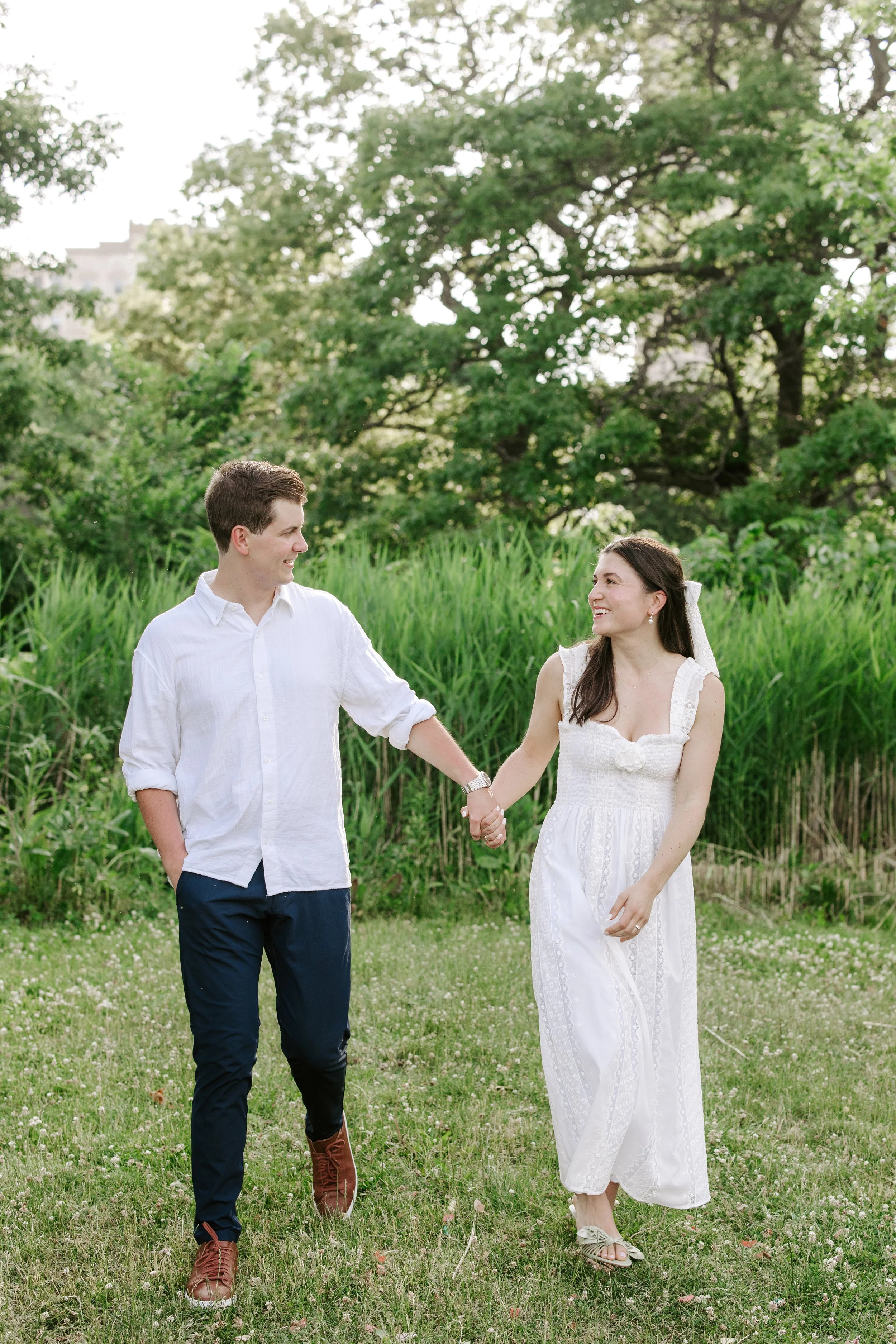 A young couple holding hands and smiling outdoors on a grassy field with trees in the background.