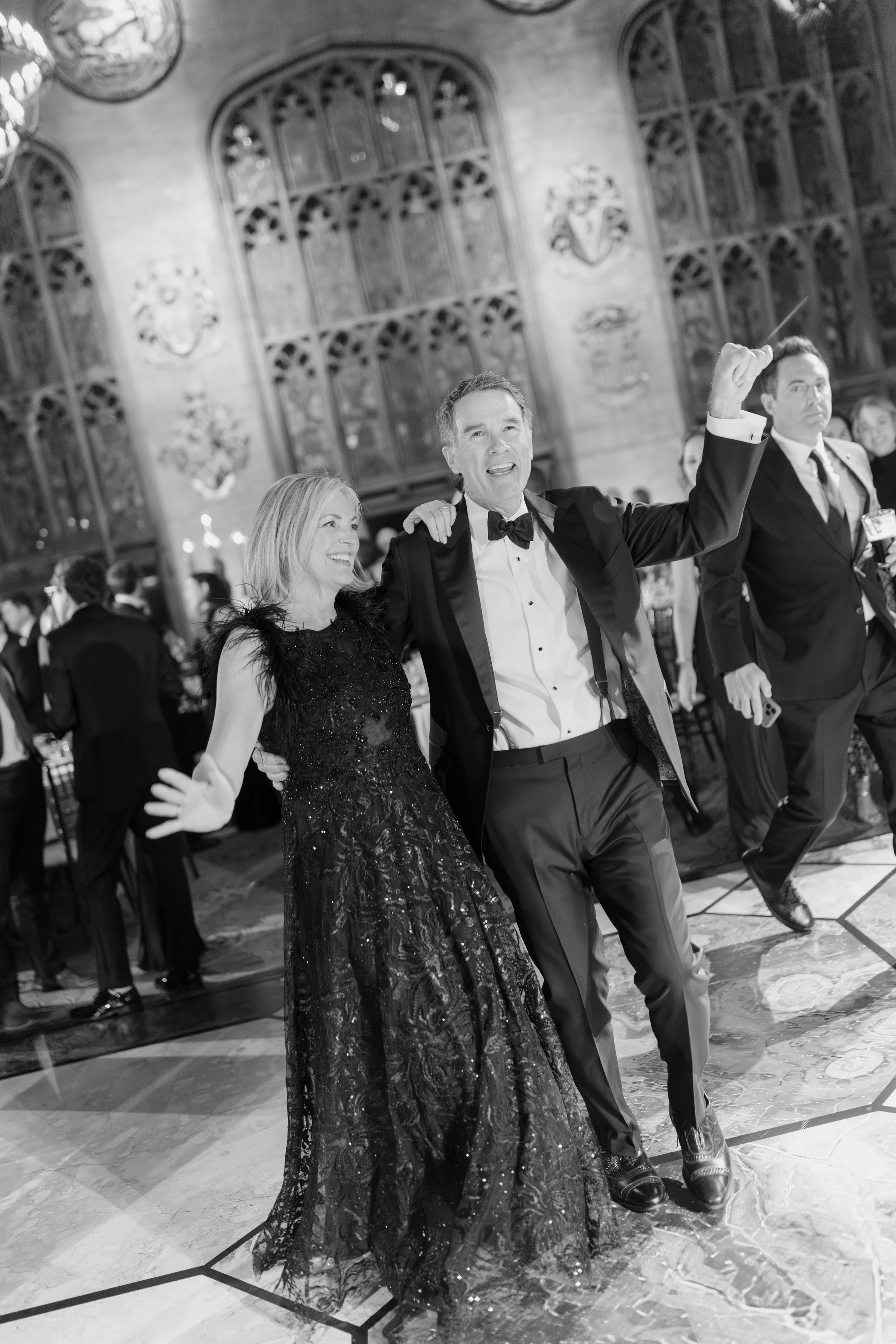 People dancing and celebrating at a formal event in a grand hall with stained glass windows. A man in a tuxedo is smiling and raising his arm, accompanied by a woman in an elegant black dress. Other guests are visible in the background.