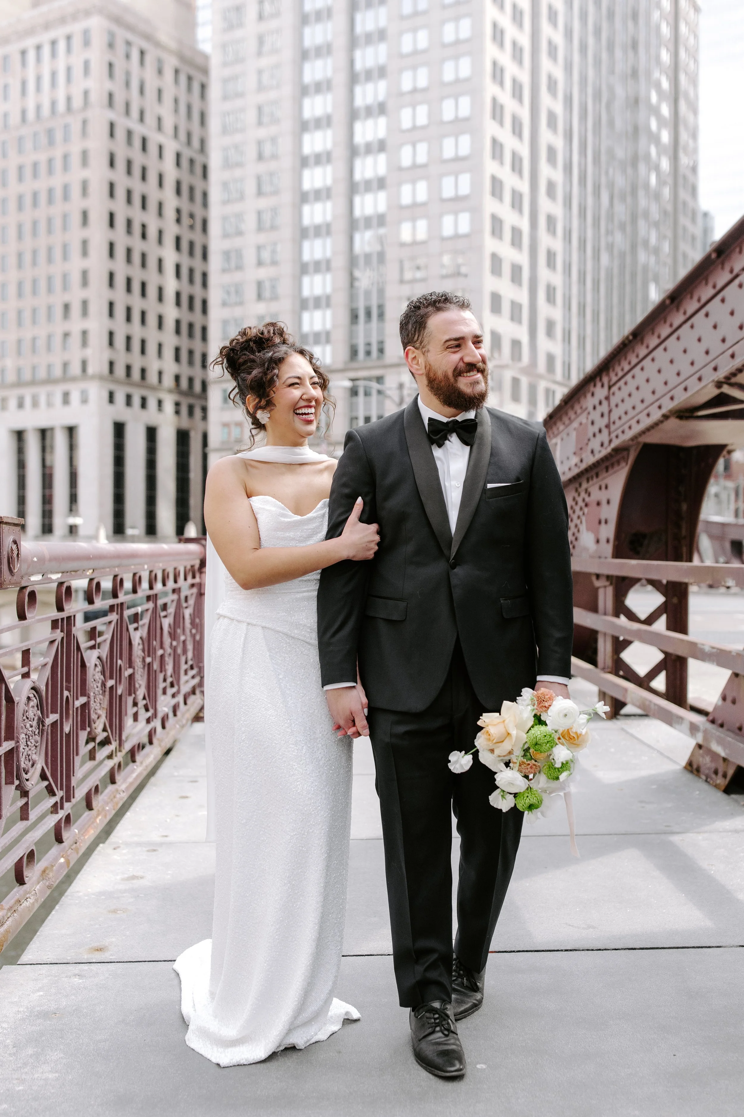 A bride and groom in wedding attire standing on a city bridge with tall buildings in the background. The bride is smiling and behind the groom, holding his arm, wearing a white wedding dress. The groom is in a black tuxedo with a bow tie, holding a b