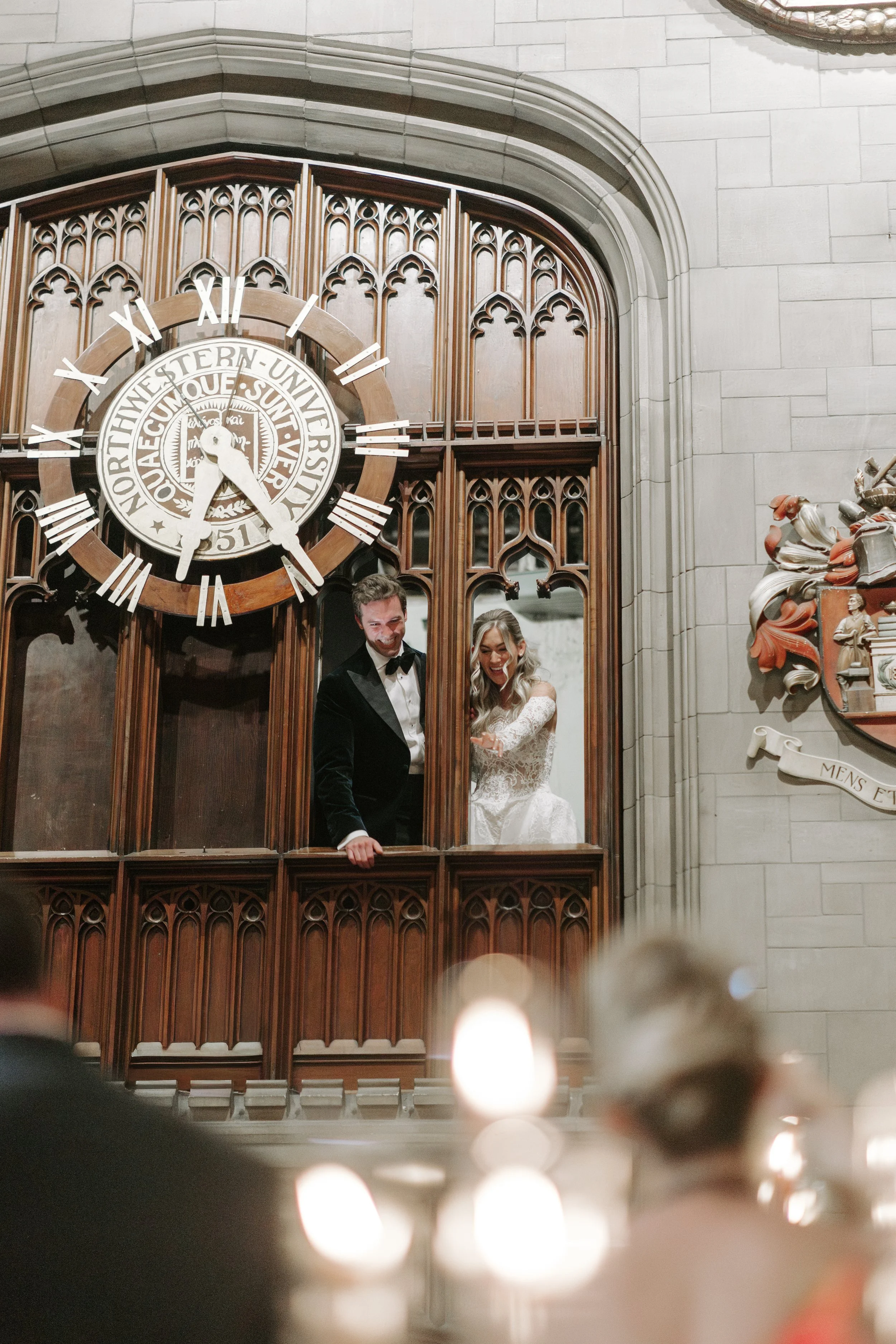 A bride and groom standing on a wooden balcony inside a church, with a large clock showing the time as approximately 3:29, and decorative Gothic architecture in the background.